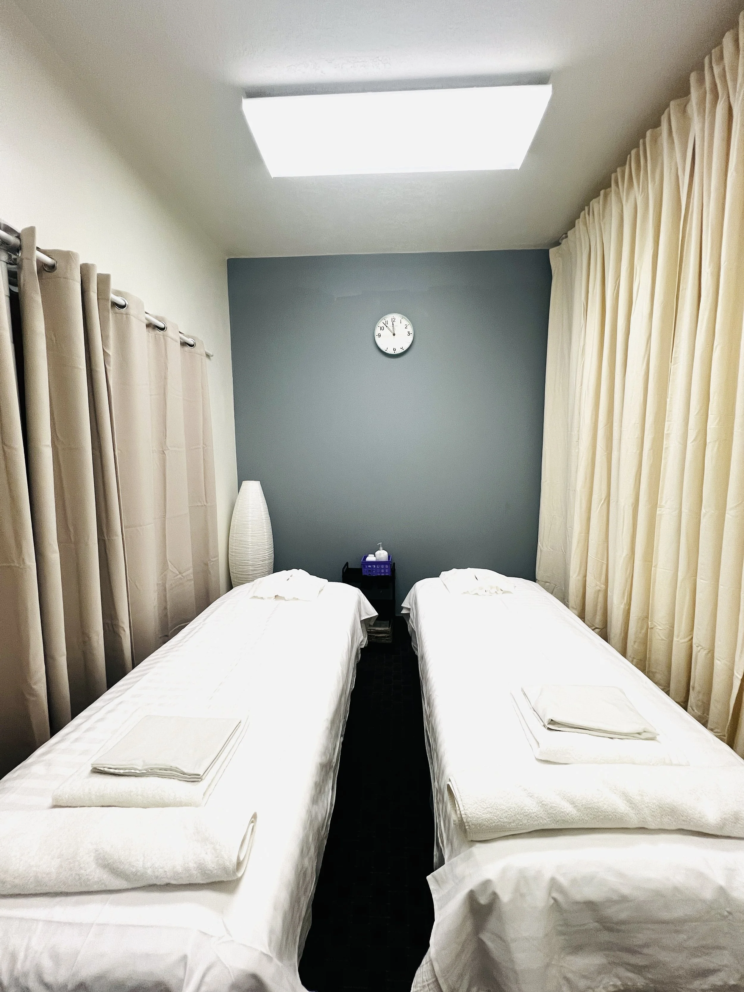 A massage table covered with white sheets and towels in a room with beige walls, beige curtains, a textured floor, a side table with a lamp, clock, and decorative plants, and a tall white vase in the corner.