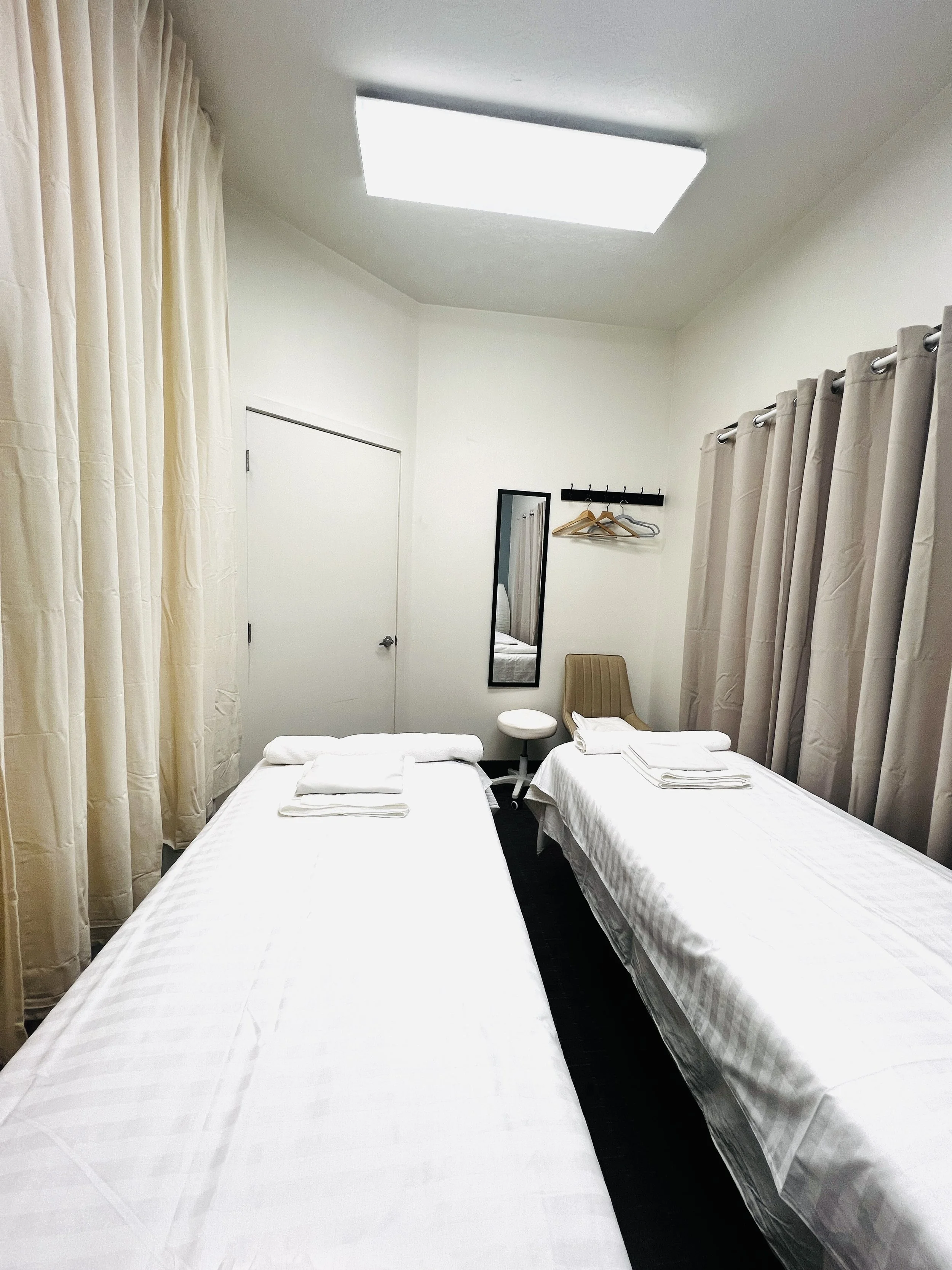 Hotel room with a bed covered in a white sheet, a small bedside table with a lamp, some toiletries, a green plant, large windows with blinds, and patterned armchairs.