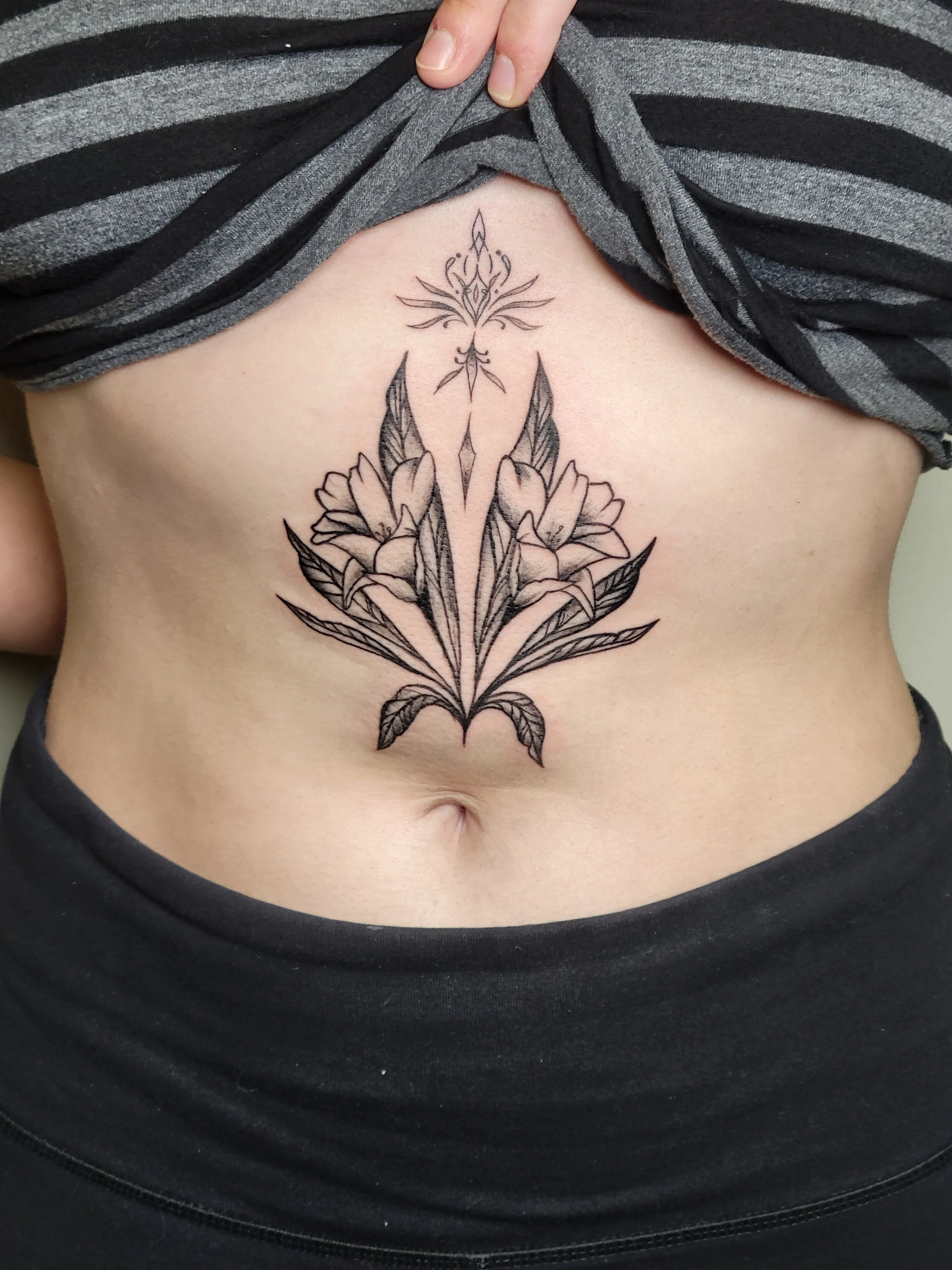 A black and gray floral tattoo on a person's stomach, featuring a large symmetrical flower with elongated leaves and an intricate decorative element above.