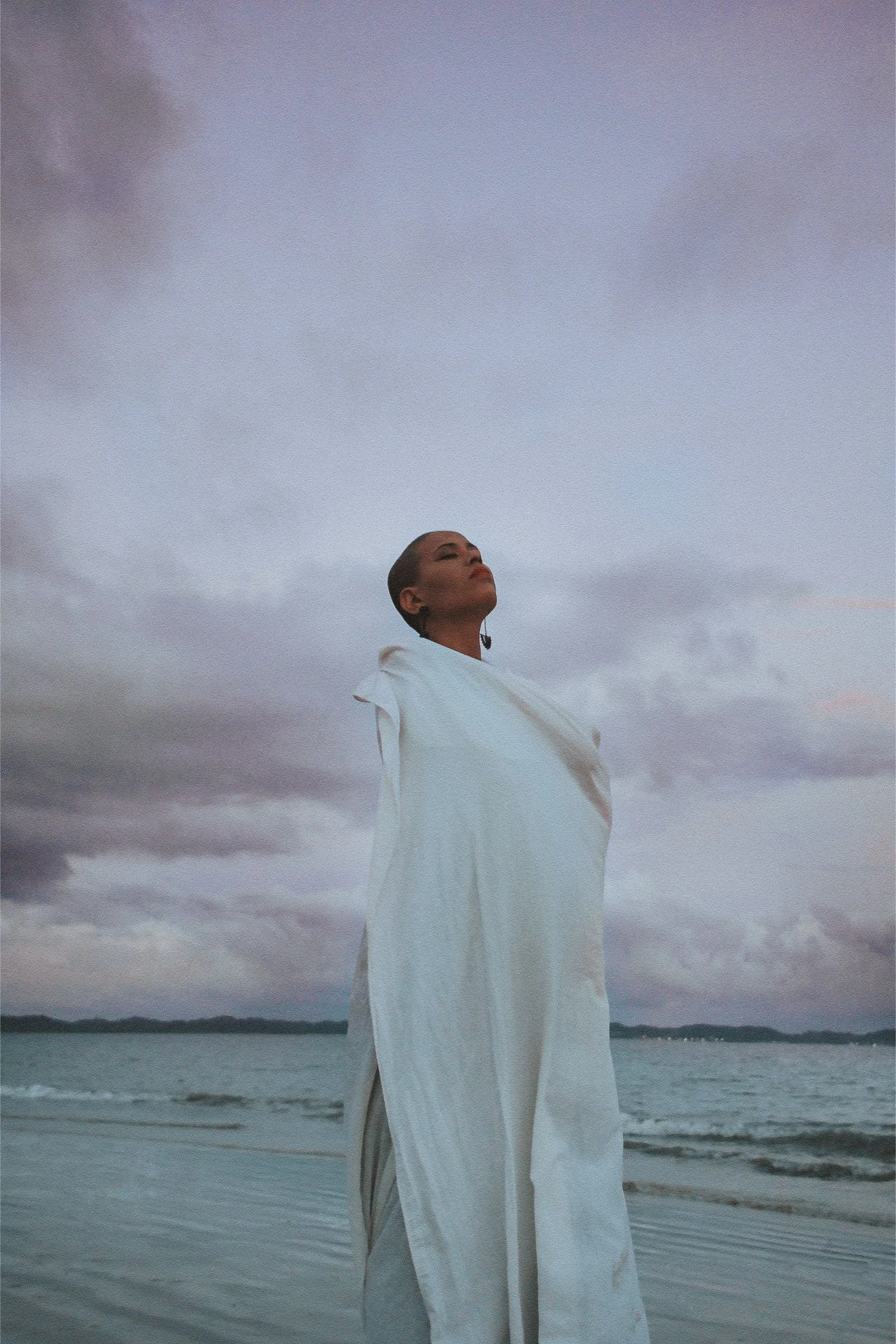 A woman standing on a beach with her eyes closed, wearing a white long dress, under a cloudy sky during sunset.