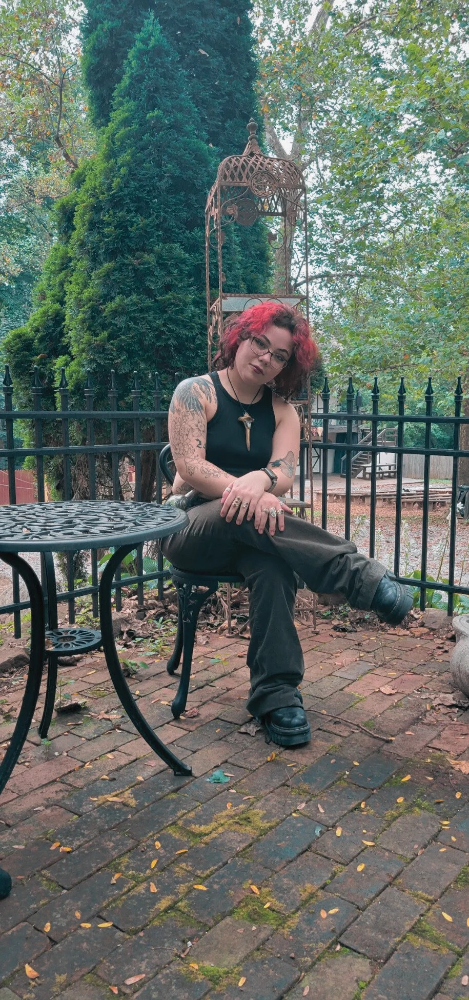 A woman with red and black curly hair, glasses, and tattoos on her arms, sitting outdoors on a black metal chair. She wears a black sleeveless top, dark pants, and black shoes, with a necklace featuring a tooth pendant. The setting includes a brick ground, a black metal fence, trees, and garden decor in the background.