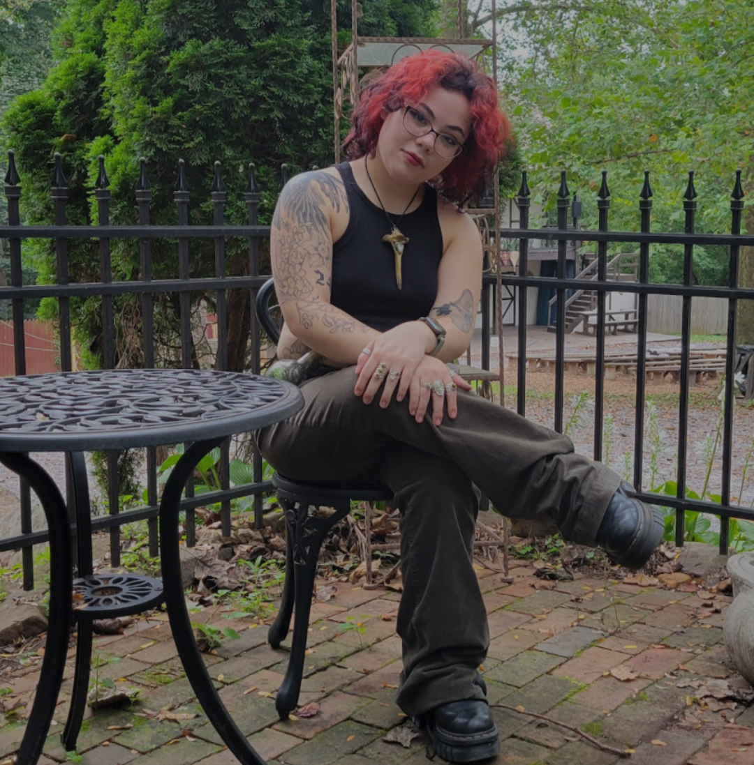 A woman with curly red hair and glasses, sitting outdoors on a patio with black metal furniture and a fence, surrounded by green trees.
