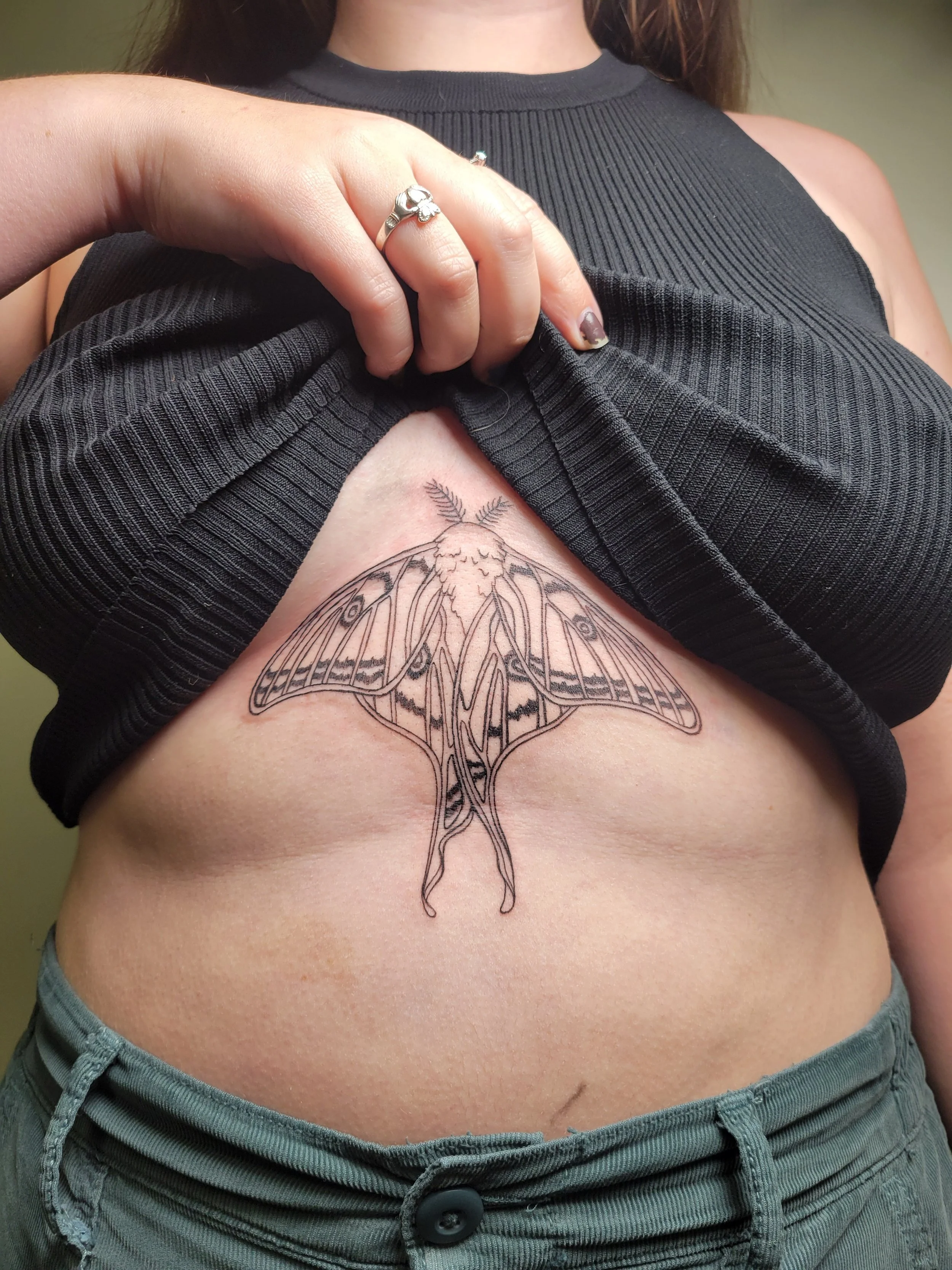 A person lifting their black ribbed tank top to reveal a tattoo of a moth on their side, just above the waist, and wearing a silver ring with a heart shape on their finger.