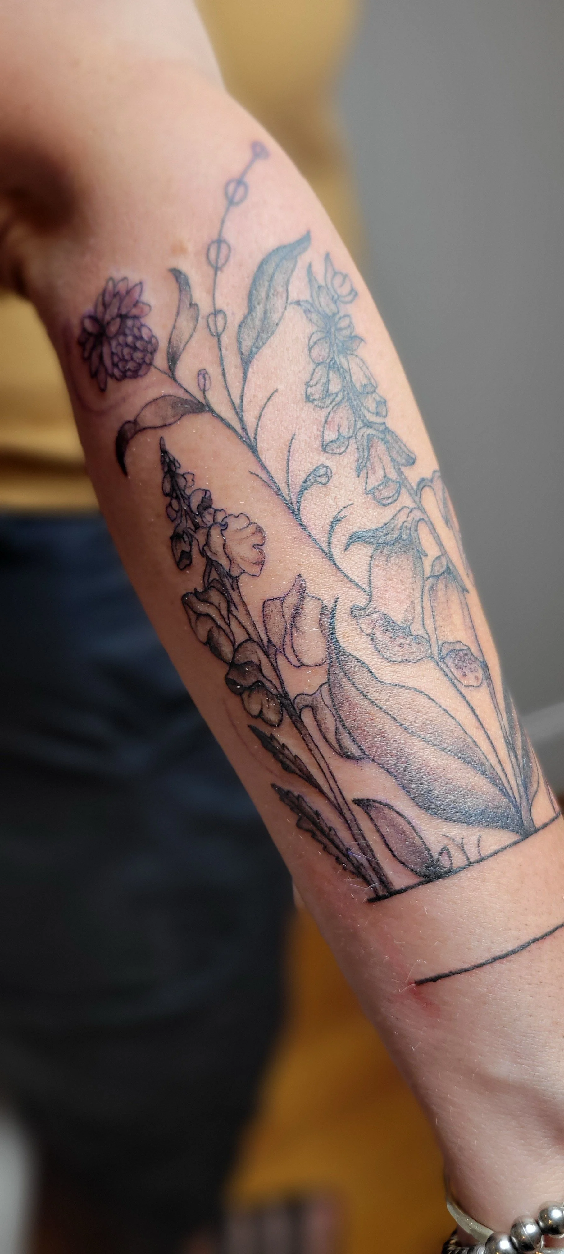 A person’s forearm with a tattoo outline of various flowers and leaves.