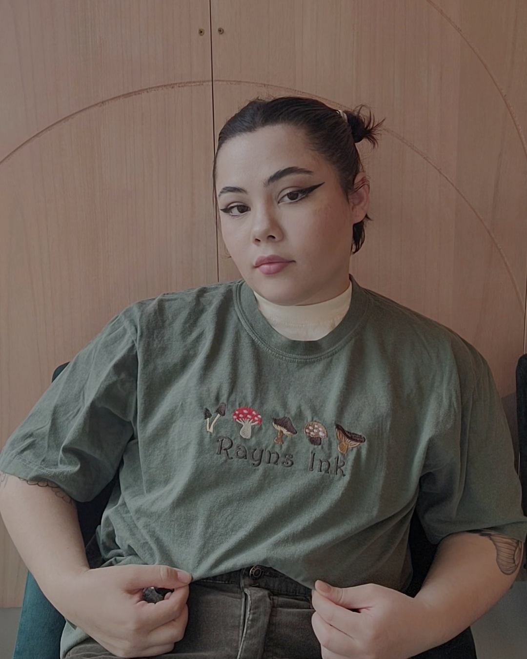 Young woman with dark hair in a bun, sitting with a confident expression, wearing a green T-shirt with mushroom embroidery and the words 'Rayns Ink' on it, in front of a wooden wall.