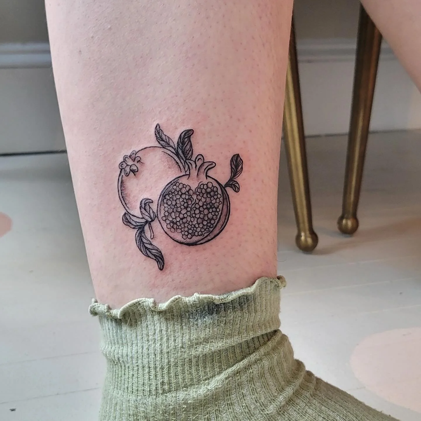 Black outline tattoo of a pomegranate with leaves on a person's calf.