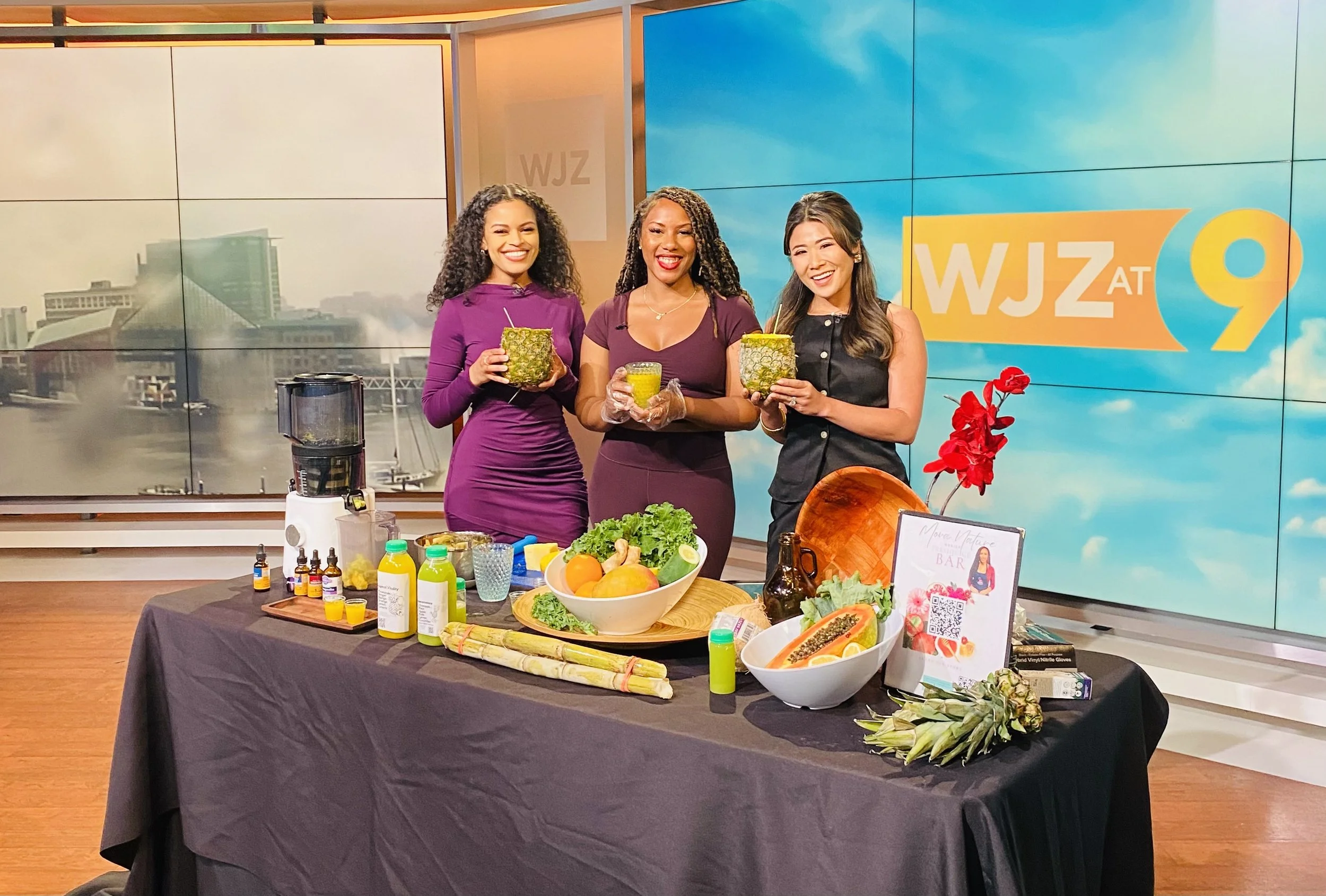 Three women standing behind a table with various fruit and health products, smiling, in a television studio with a WJZ at 9 background.