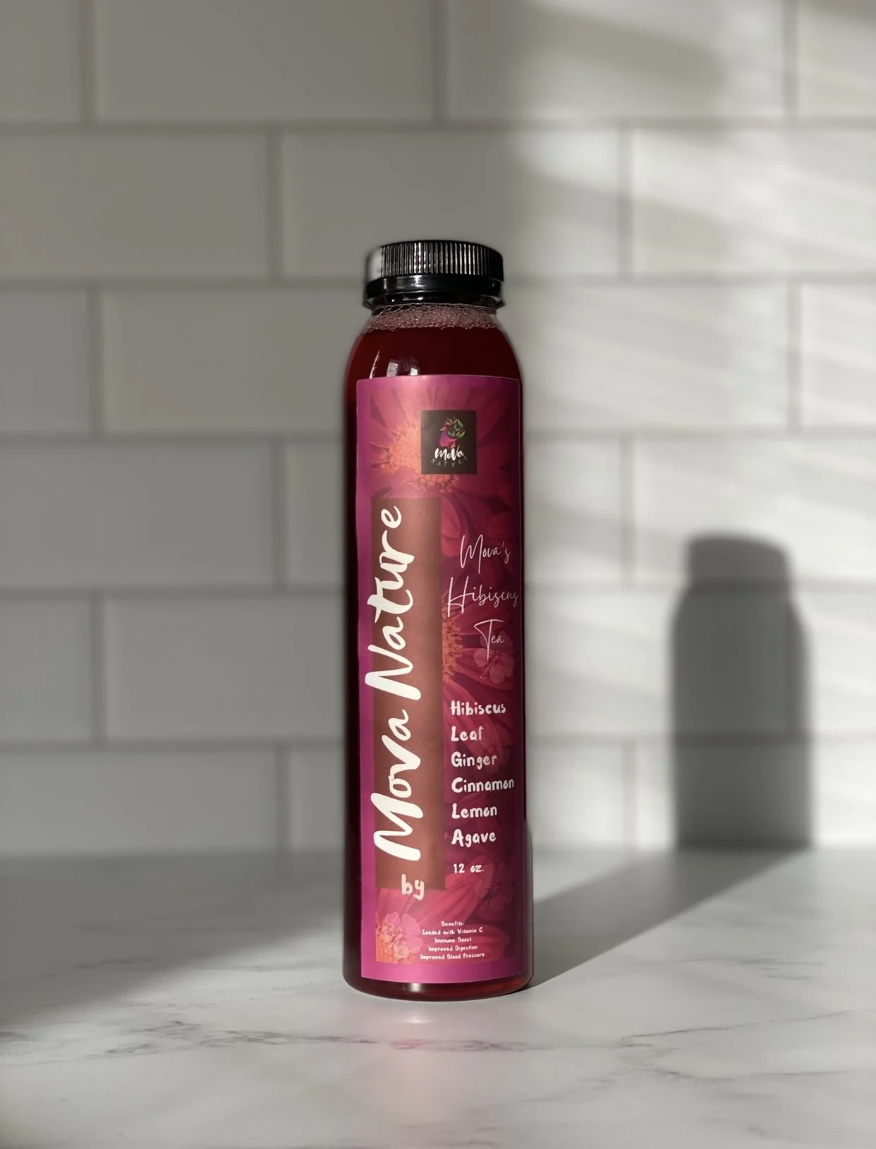 A bottle of Mona Nature herbal tea with hibiscus, leaf, ginger, cinnamon, lemon, and agave on a marble surface in front of a white tiled wall.