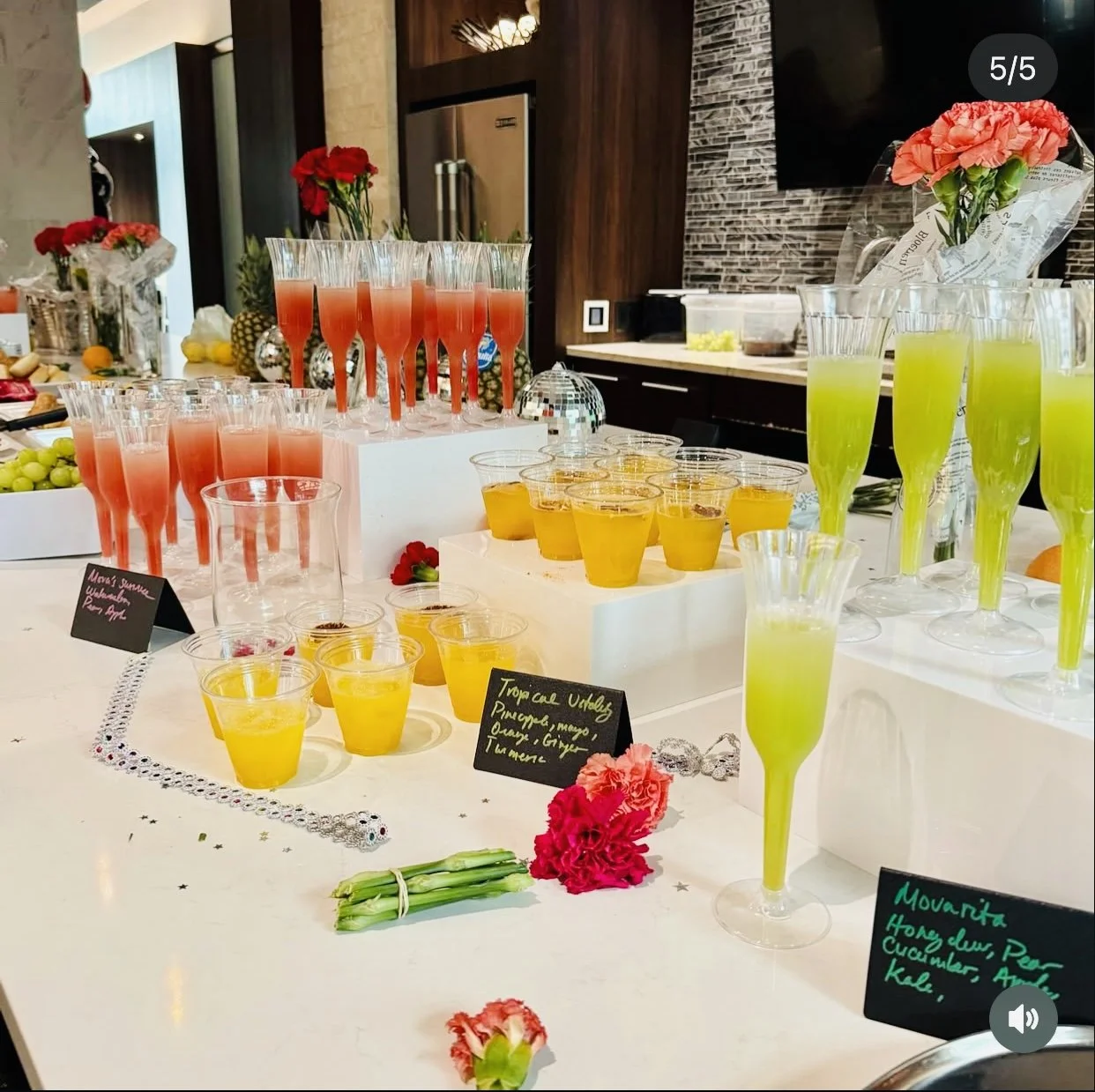 A colorful array of beverages on a white table, including pink, yellow, and green drinks in various glasses, with flowers, fruits, and decorative signs at a party or event setting.