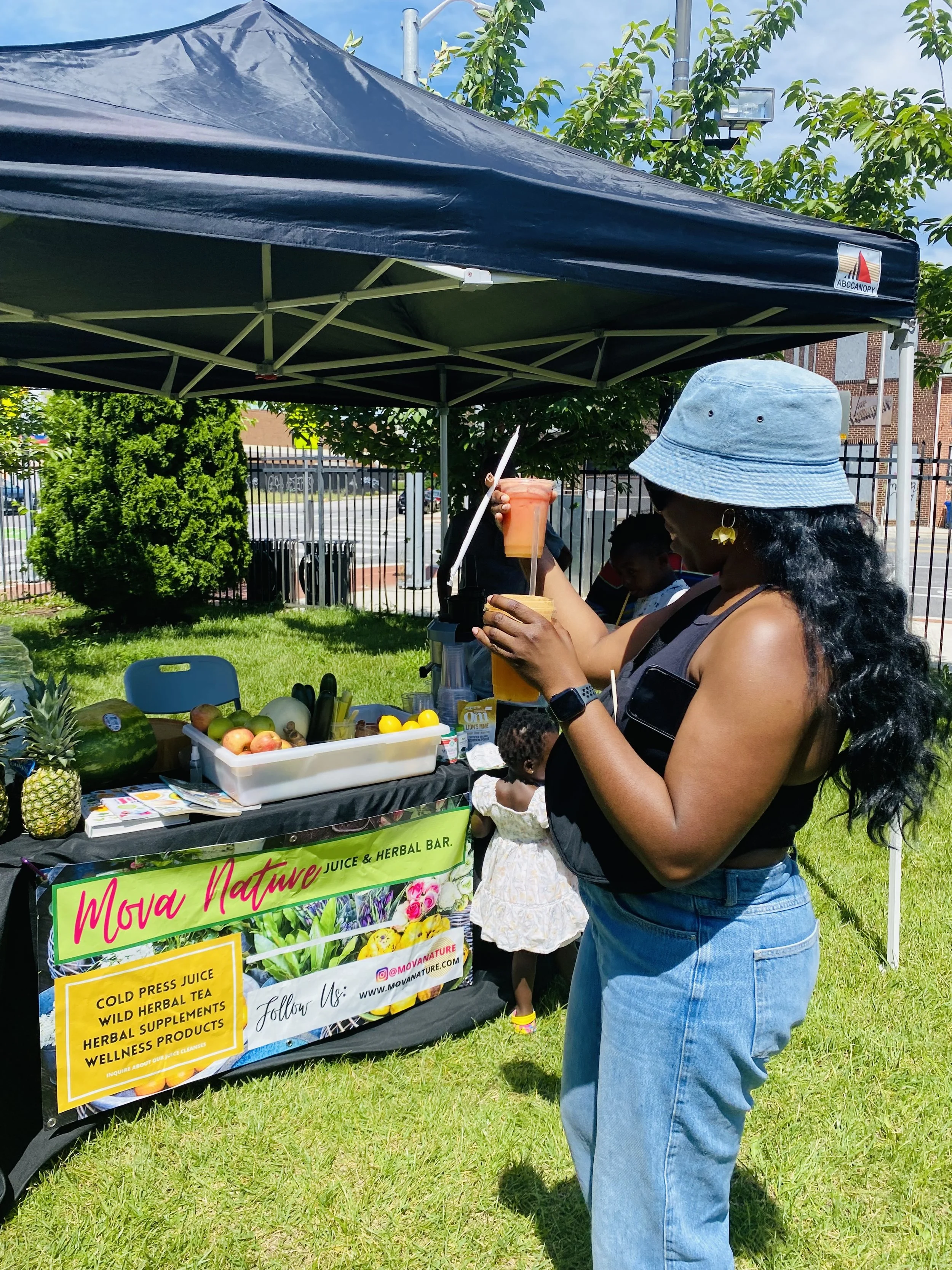 A woman in a denim hat and sleeveless top at an outdoor juice stand, holding a pink smoothie with a straw and a yellow drink, with a small child in a white dress nearby, a table with fruits and a sign that reads 'Mova Nature Juice & Herbal Bar.'