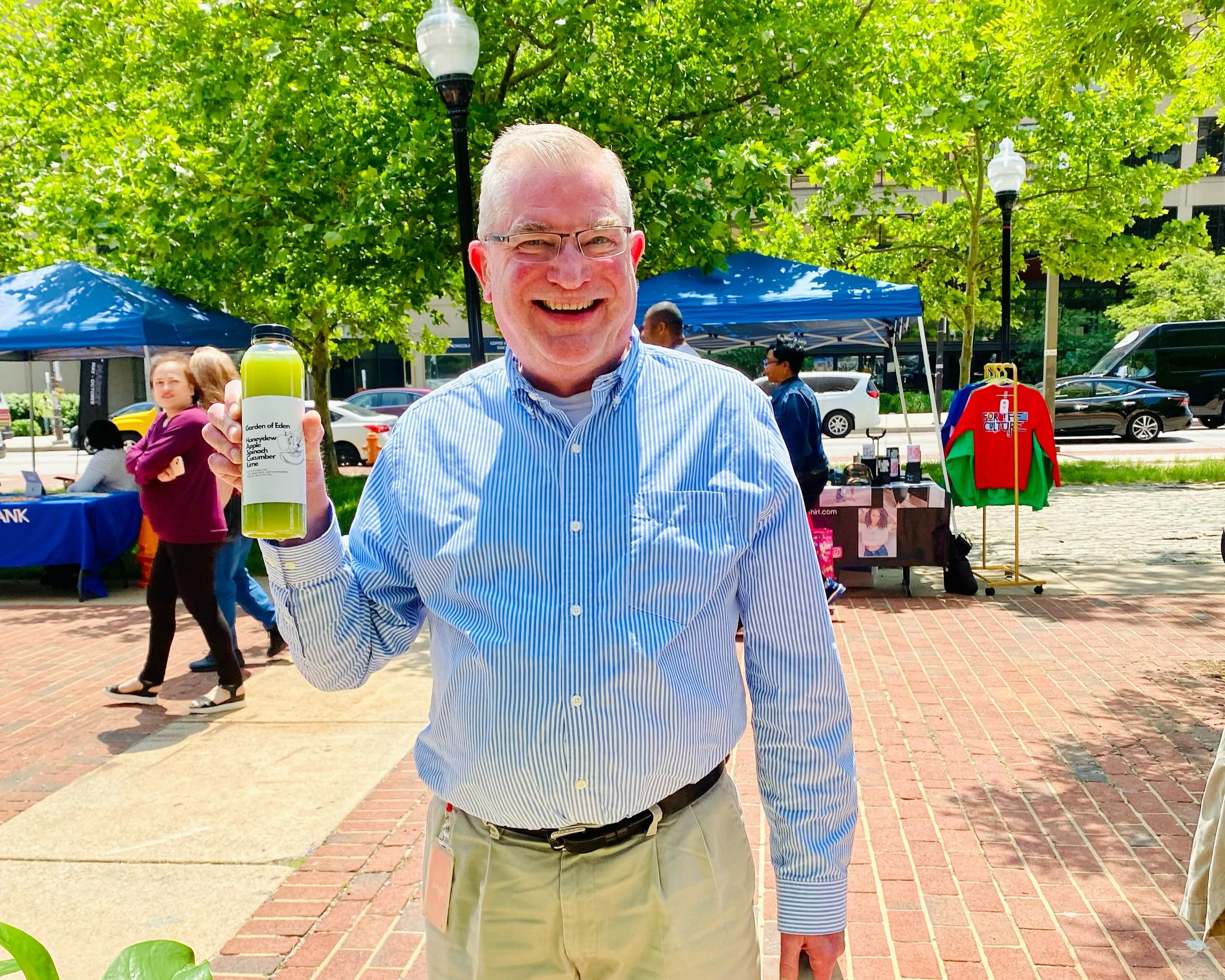 A smiling man in a blue striped shirt holding a green bottle at an outdoor event with trees, tents, and people in the background.