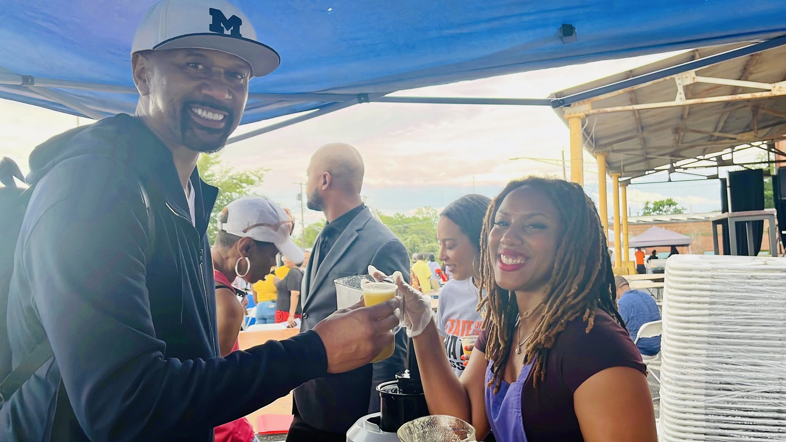 A man with a cap smiling and holding a drink, standing beside a woman with dreadlocks smiling, at an outdoor event under a tent with other attendees in the background.