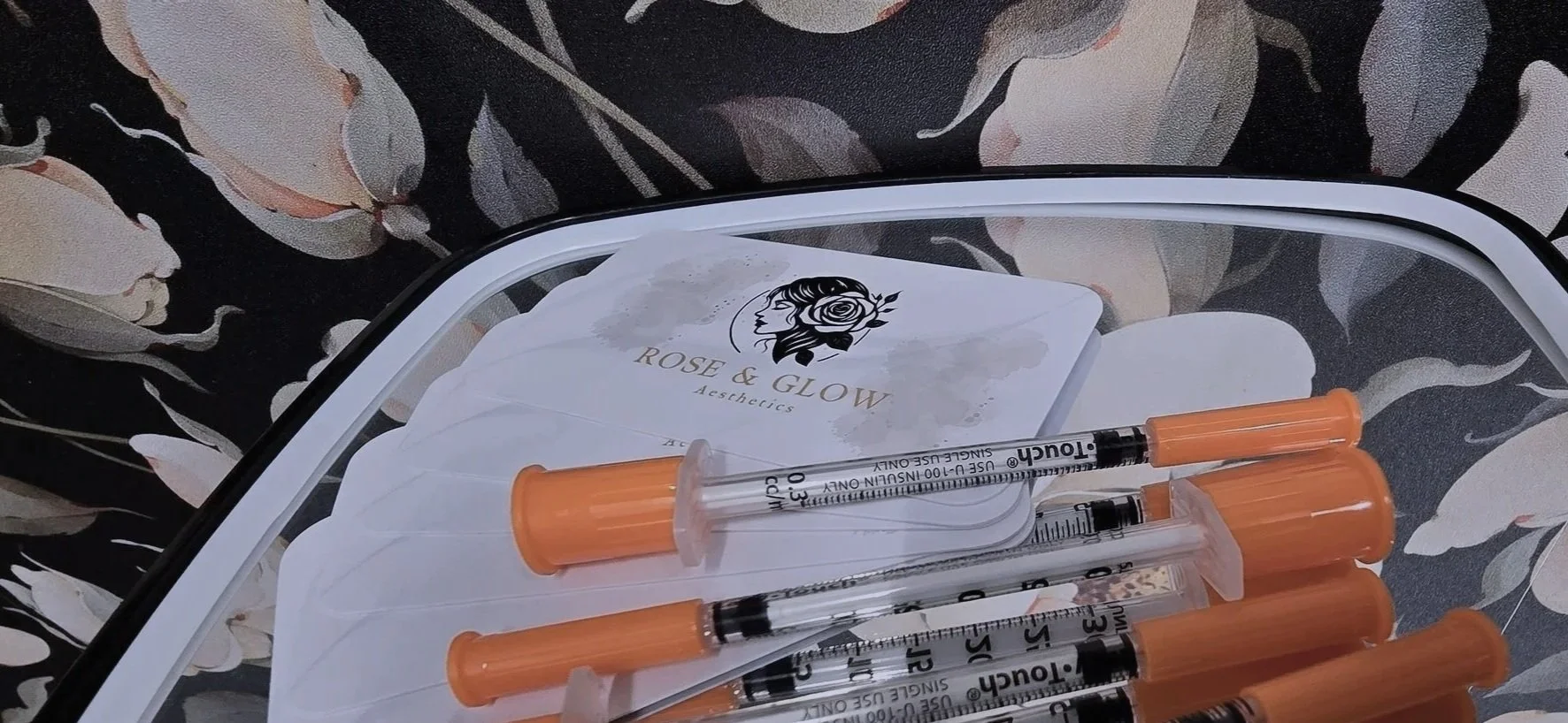 Medical syringes with orange caps resting on cosmetic product brochures labeled 'Rose & Glow Aesthetics' on a reflective surface with floral patterned background.