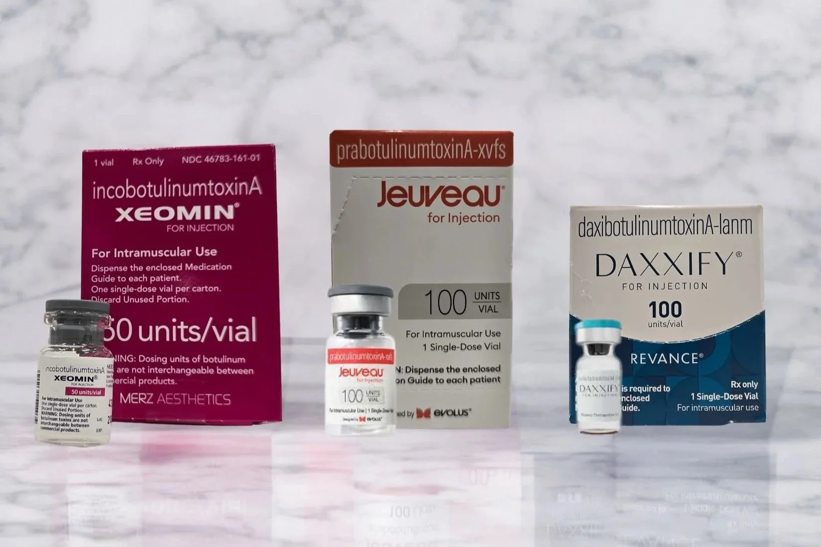 Three vials of botulinum toxin medications with packaging in the background. The boxes are purple for Xeomin, white and red for Jeuveau, and white and blue for Daxxify.