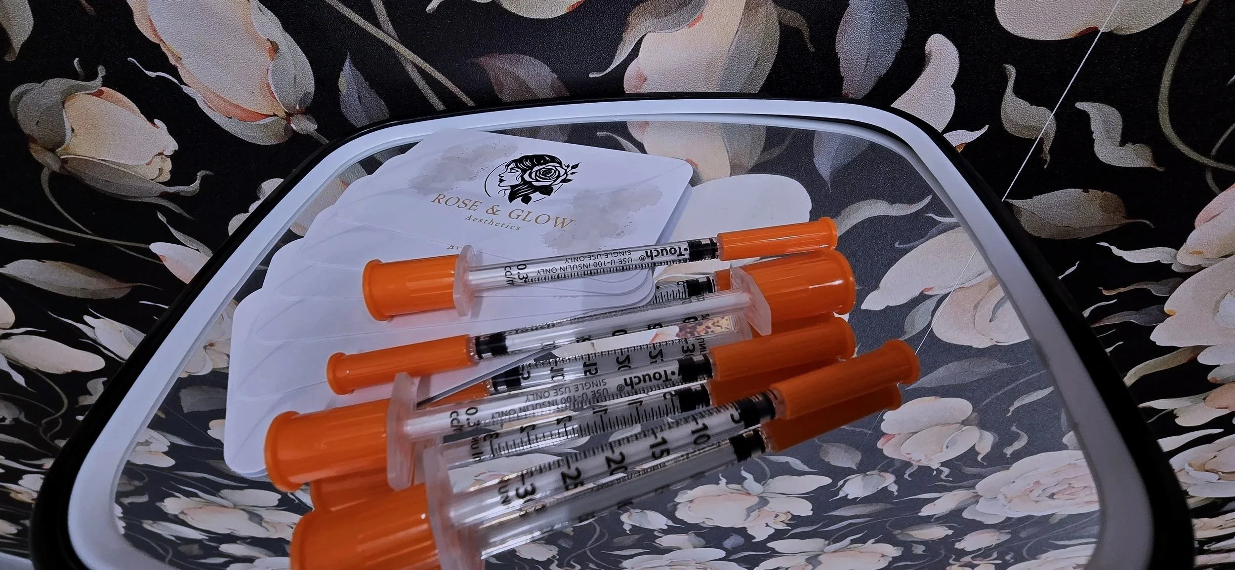 Six syringes with orange caps and a stack of facial masks with a logo that reads 'Rose & Glow Aesthetics' in front of a floral-patterned background.