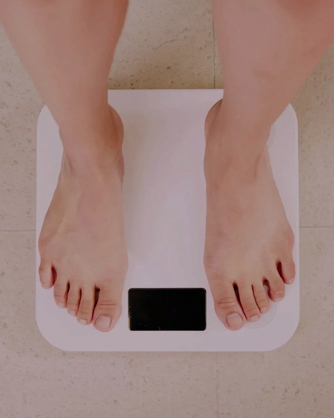 Top-down view of a person's feet on a white digital scale, with a black display at the front, on a beige tiled floor.
