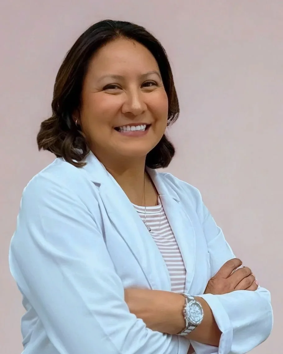 A smiling woman wearing a white lab coat, crossed arms, with a striped shirt underneath and a silver watch, standing against a plain light-colored background.