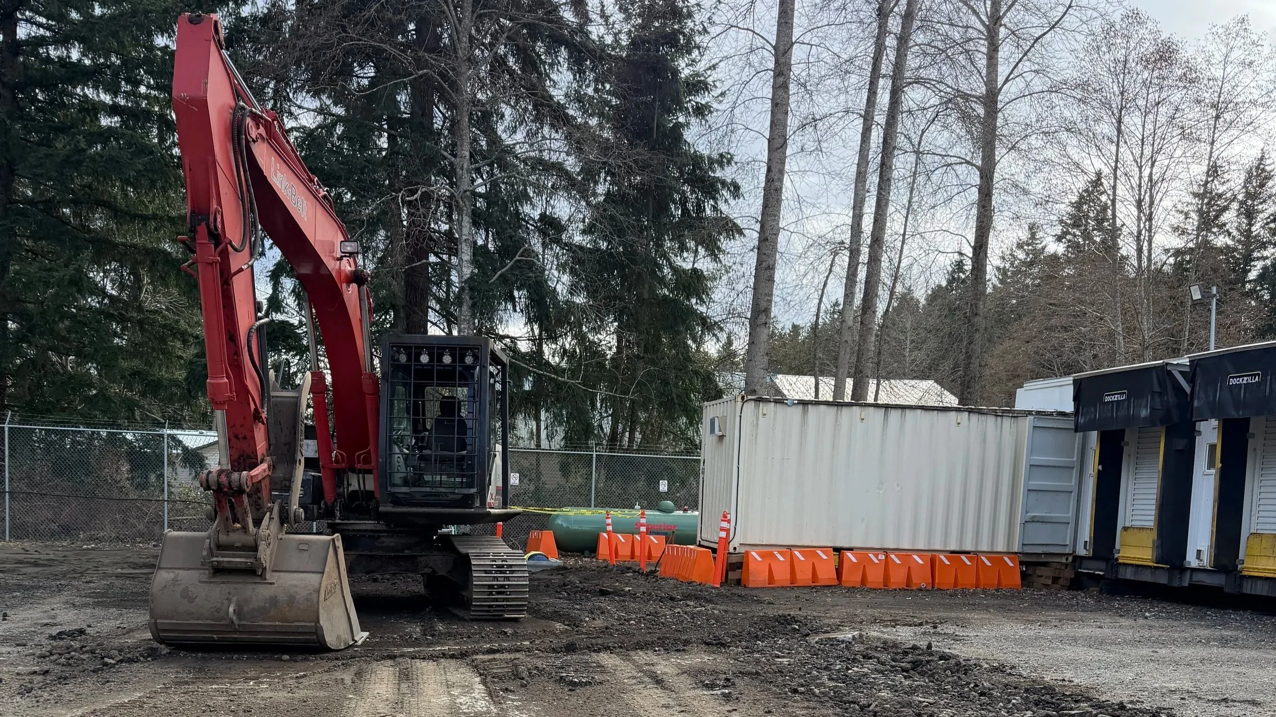 Construction site with an excavator, orange traffic cones, and portable storage units, surrounded by trees and a chain-link fence.