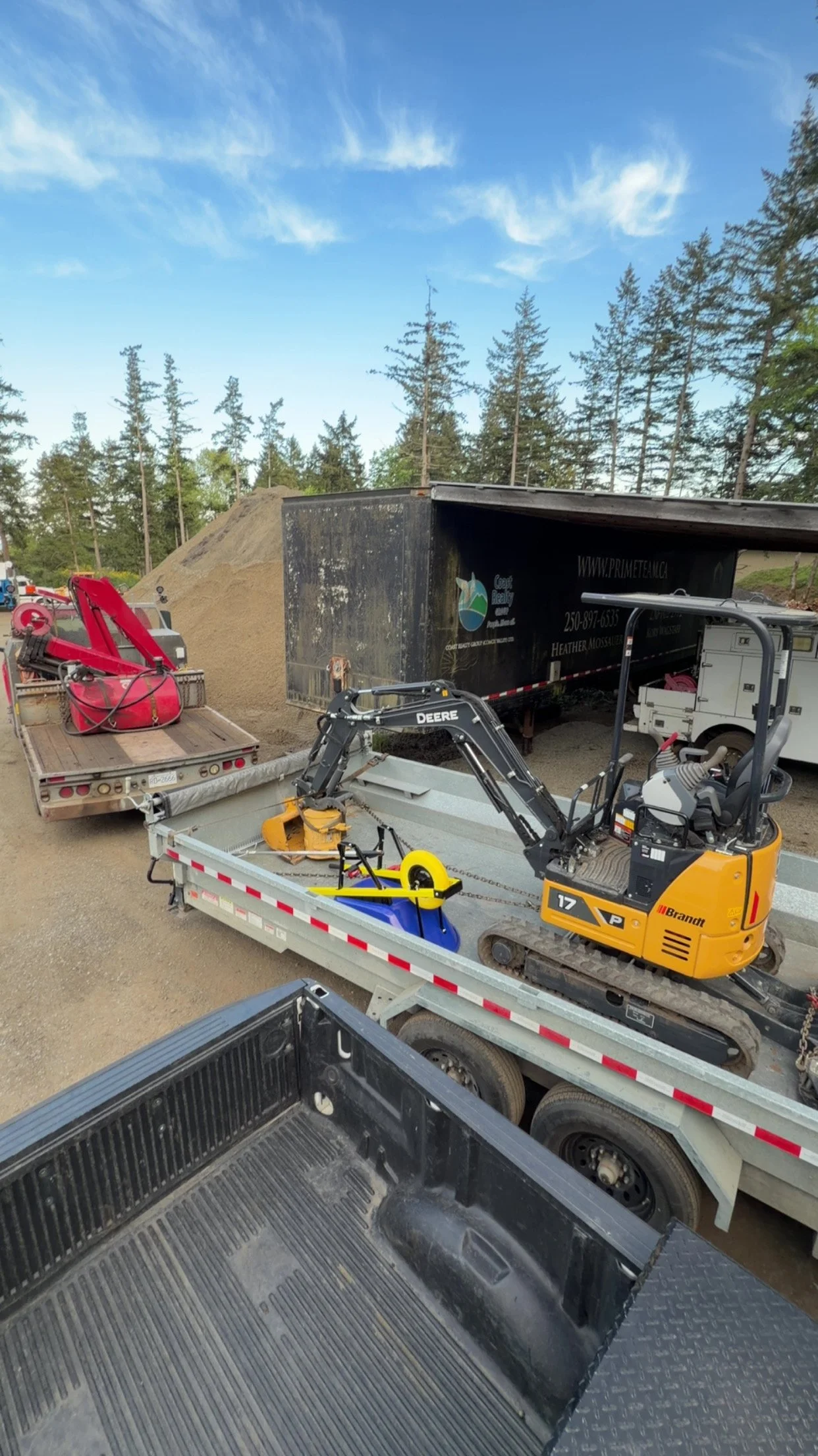 A construction site with a large pile of gravel, a small yellow excavator, and various trucks and equipment under a blue sky with some clouds, surrounded by evergreen trees.