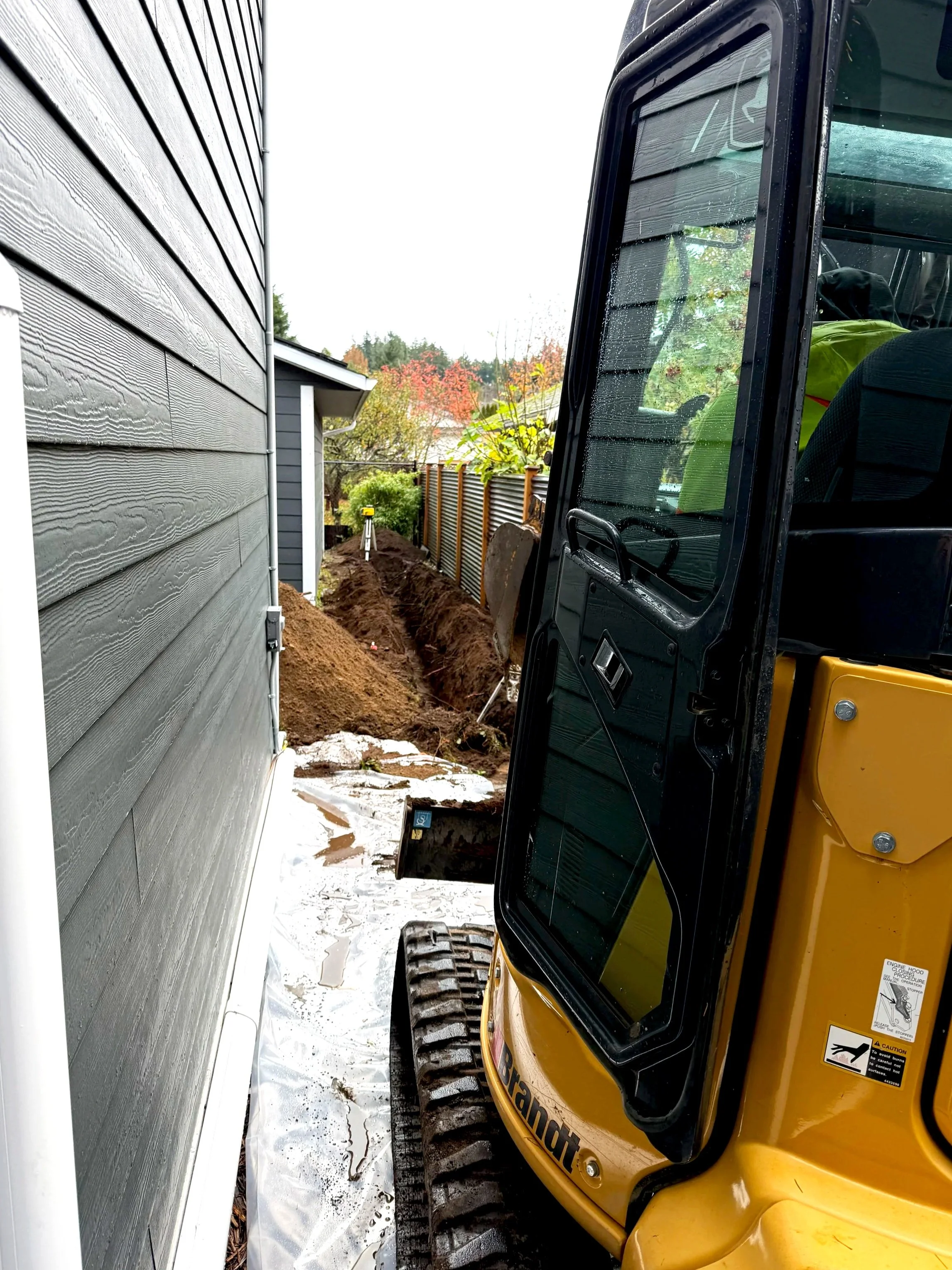 Construction or excavation machinery parked in a narrow alleyway beside a house with gray siding. The ground shows clear signs of recent digging, with dirt and debris present.
