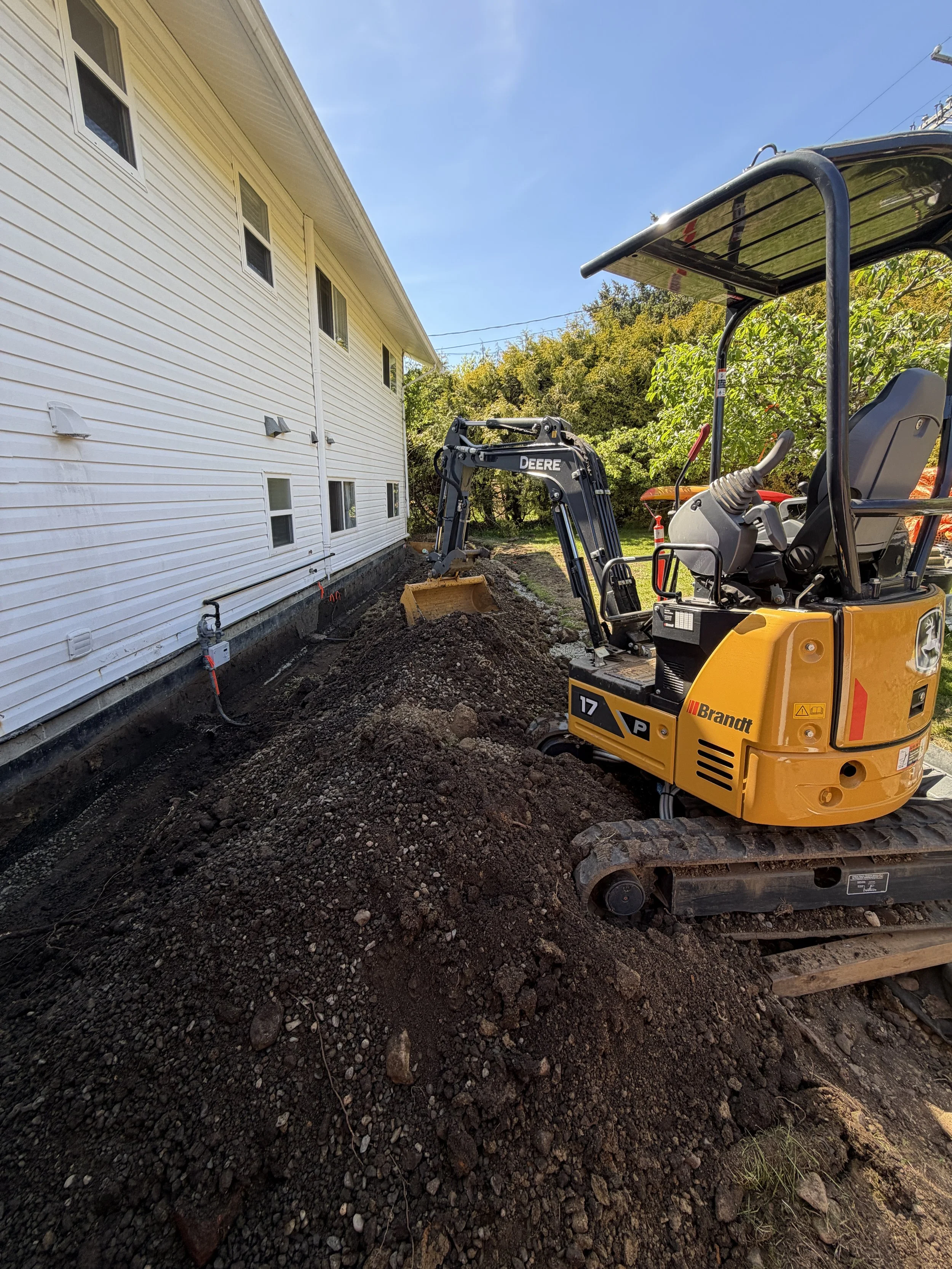 Construction site with a small yellow excavator digging a trench near a white house with beige siding and multiple small windows on an outdoor setting with trees and blue sky.