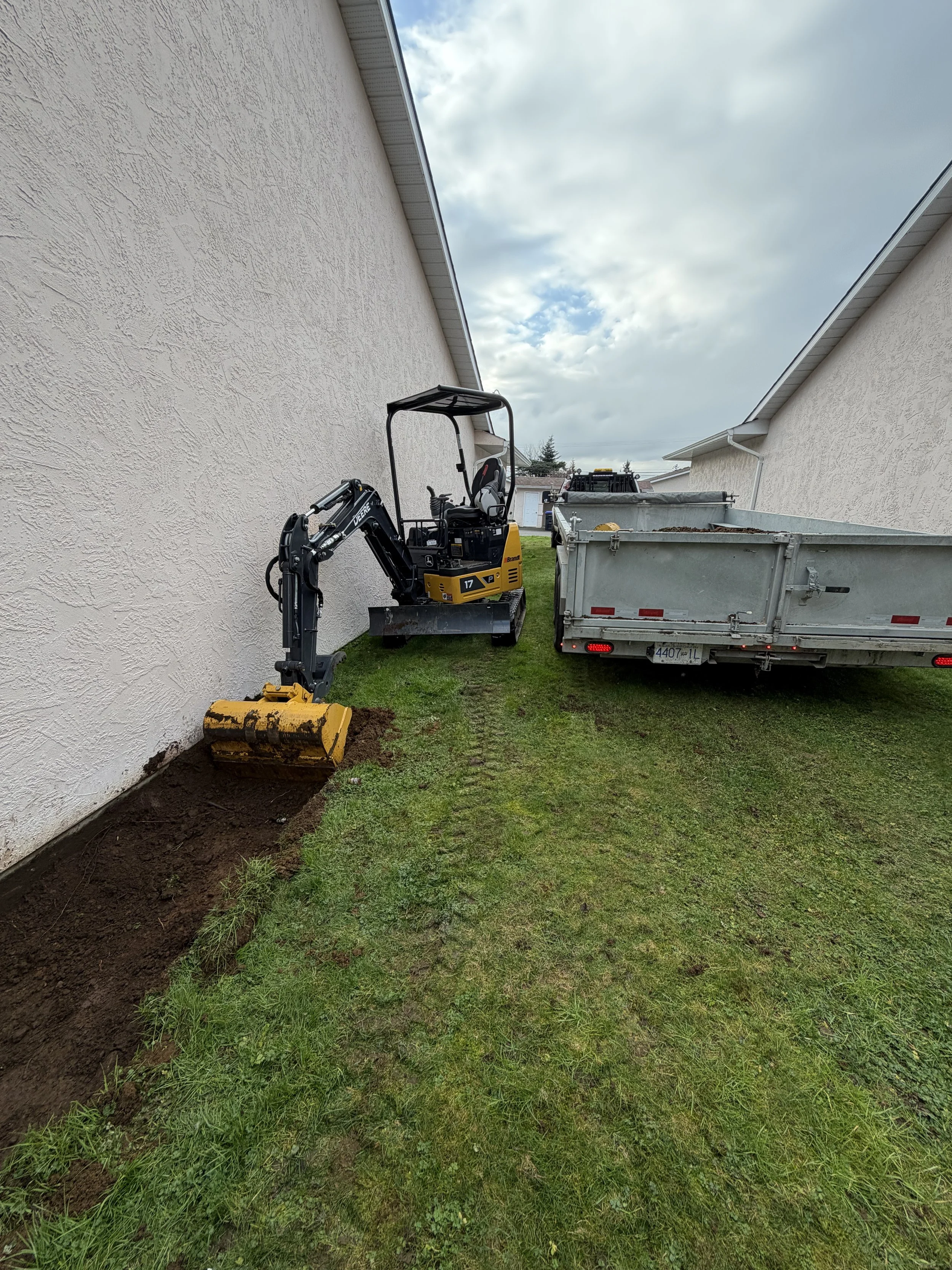 A small yellow and black excavator digging a trench along the side of a house, with a trailer parked nearby and cloudy sky overhead.