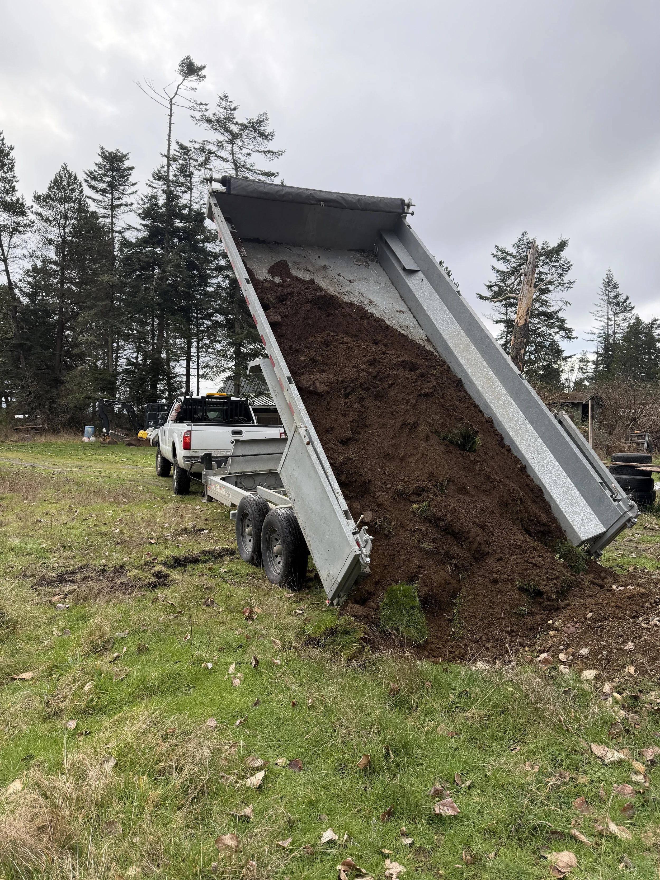 A dump truck is unloading soil onto a grassy area with trees in the background. The soil is flowing out of the back of the truck bed, creating a pile on the ground.