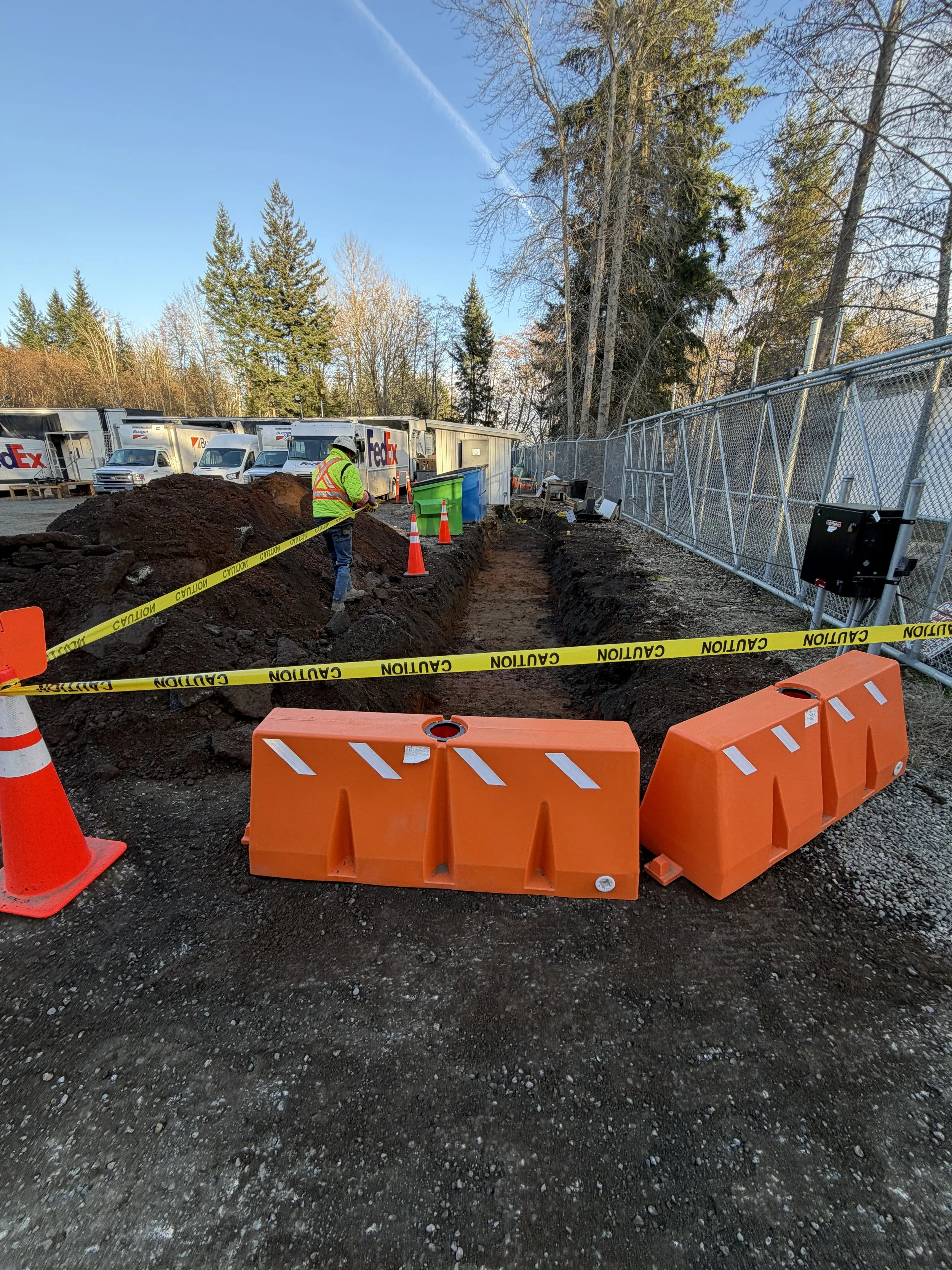 Construction site with dug trench, orange barriers, caution tape, worker in safety gear, and parked FedEx trucks in background under a partly cloudy sky.