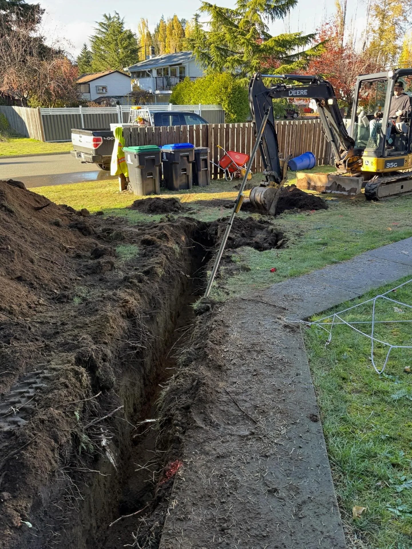 A construction site at the side of a residential street with a deep trench being dug by a small excavator. There are garbage bins lined up, a wheelbarrow, and a blue barrel, with houses and trees in the background.