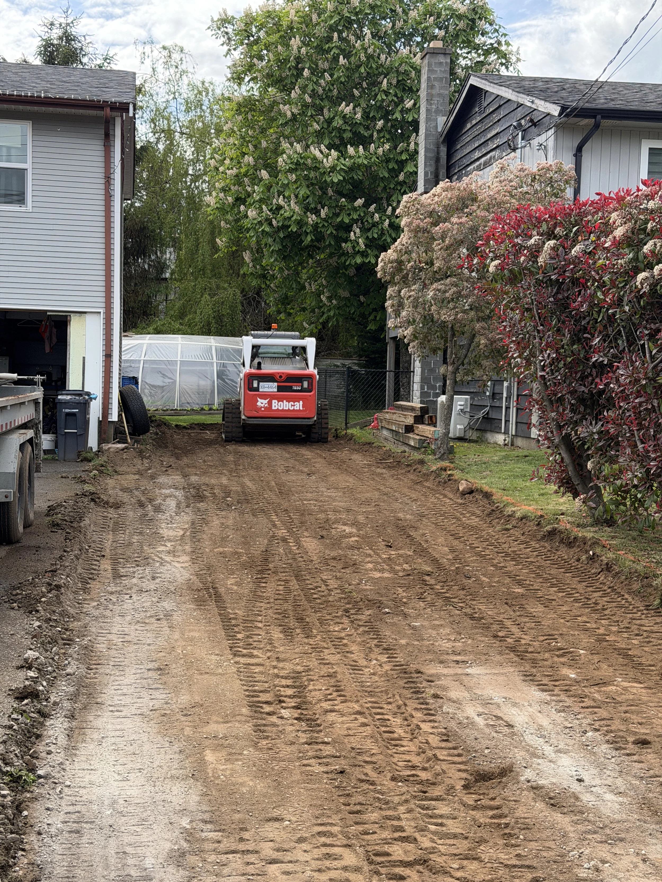 A dirt driveway with tire tracks leading to a small red Bobcat construction machine, with a neighborhood house on the right and trees in the background.