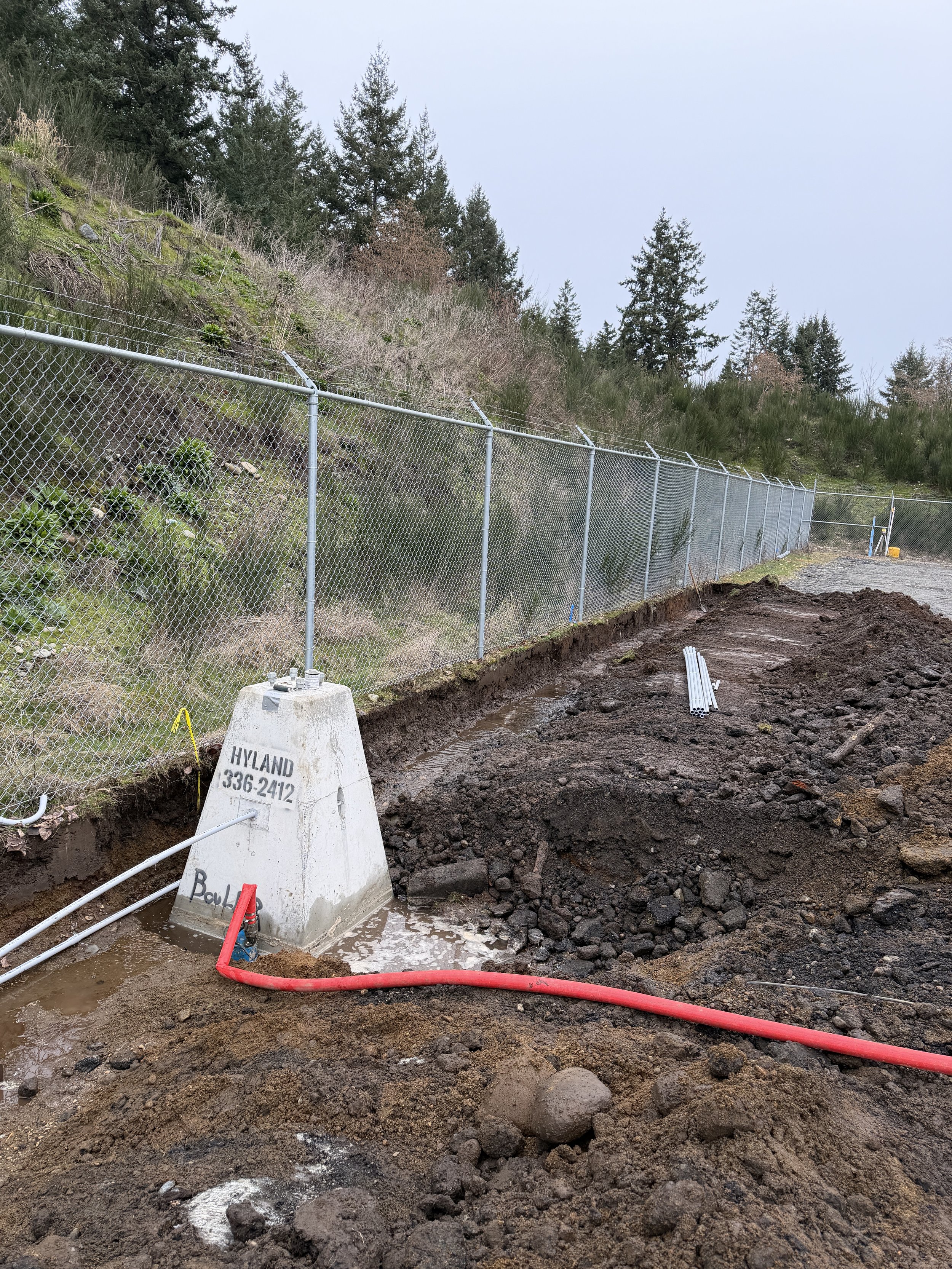 Construction site with a chain-link fence along a hillside, soil excavation, orange hoses, and a concrete marker labeled 'HYLAND 336-2412' in a mountainous area with trees.