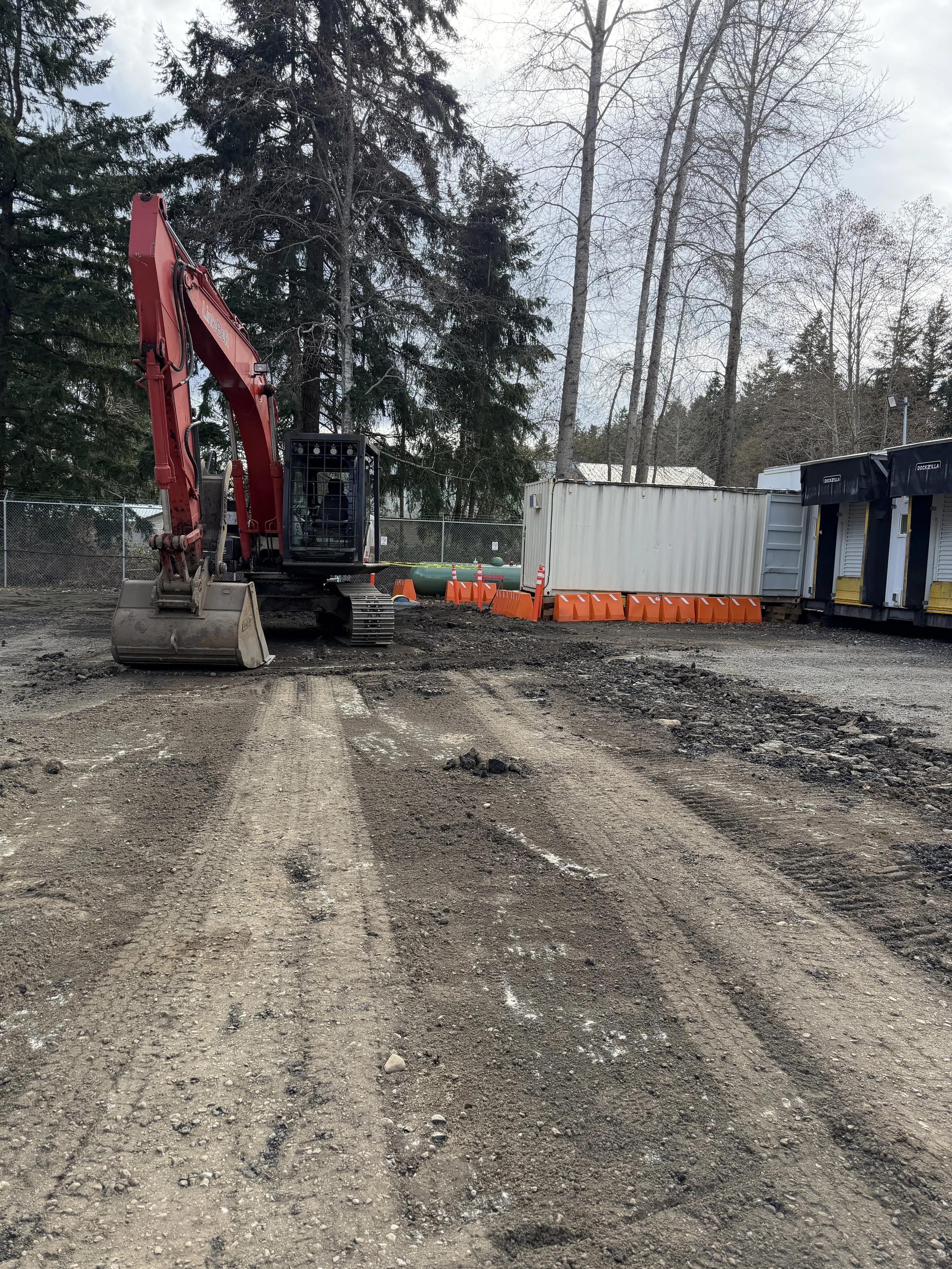 A construction site with a red excavator, orange barriers, and portable storage units, surrounded by tall trees and a chain-link fence.