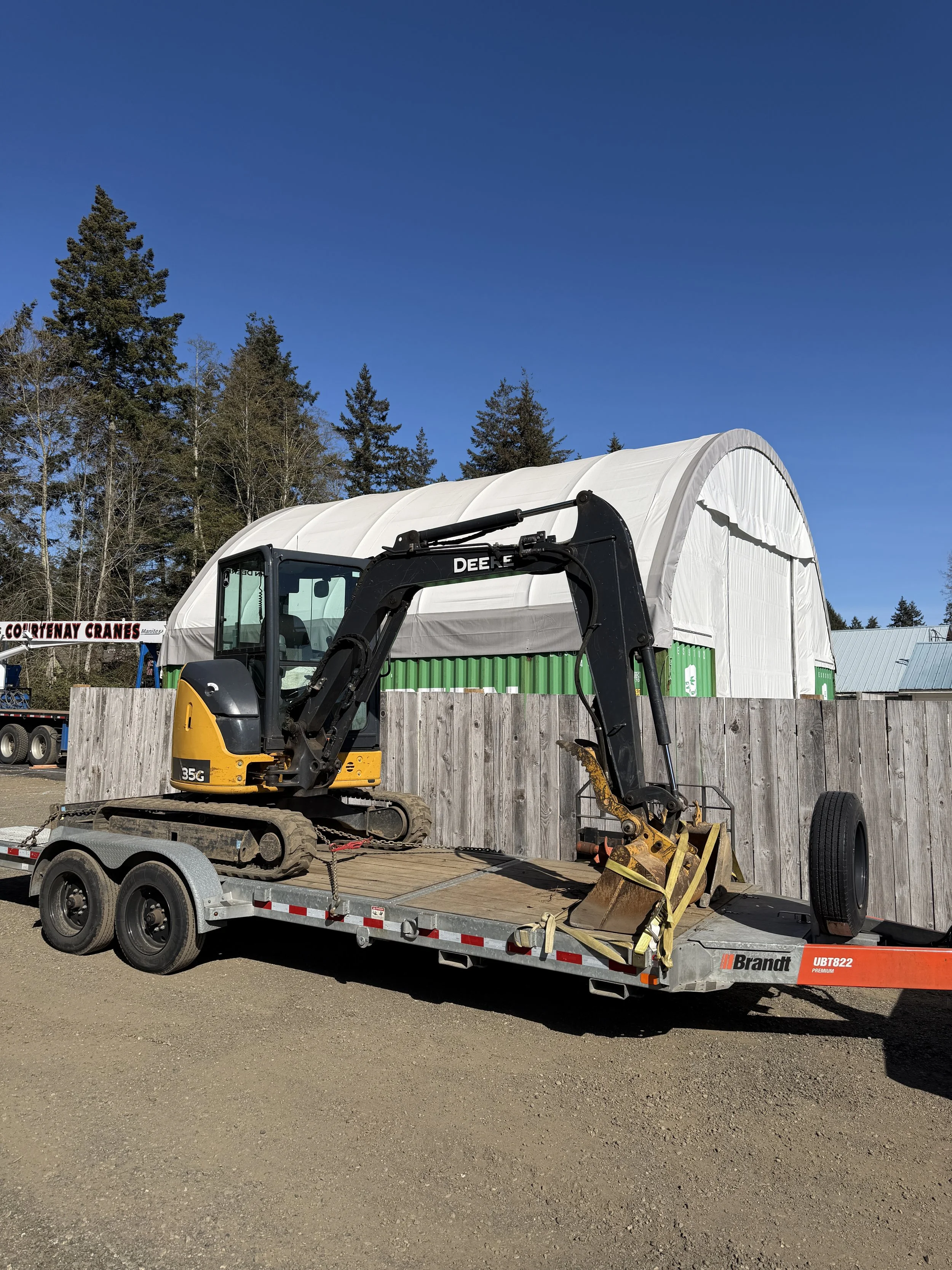 A small yellow and black excavator on a flatbed trailer, with a large white tent structure and green storage containers in the background, trees and a blue sky overhead.