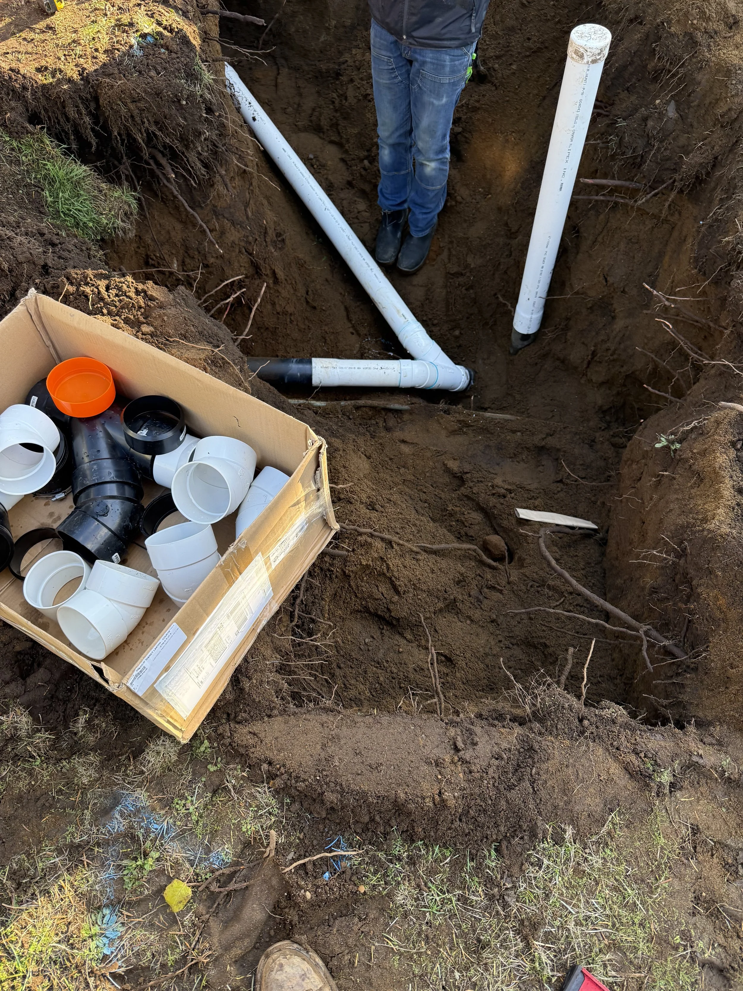 A person on a construction site installing underground piping in a deep trench surrounded by dirt and soil, with PVC pipes, fittings, and tools nearby.