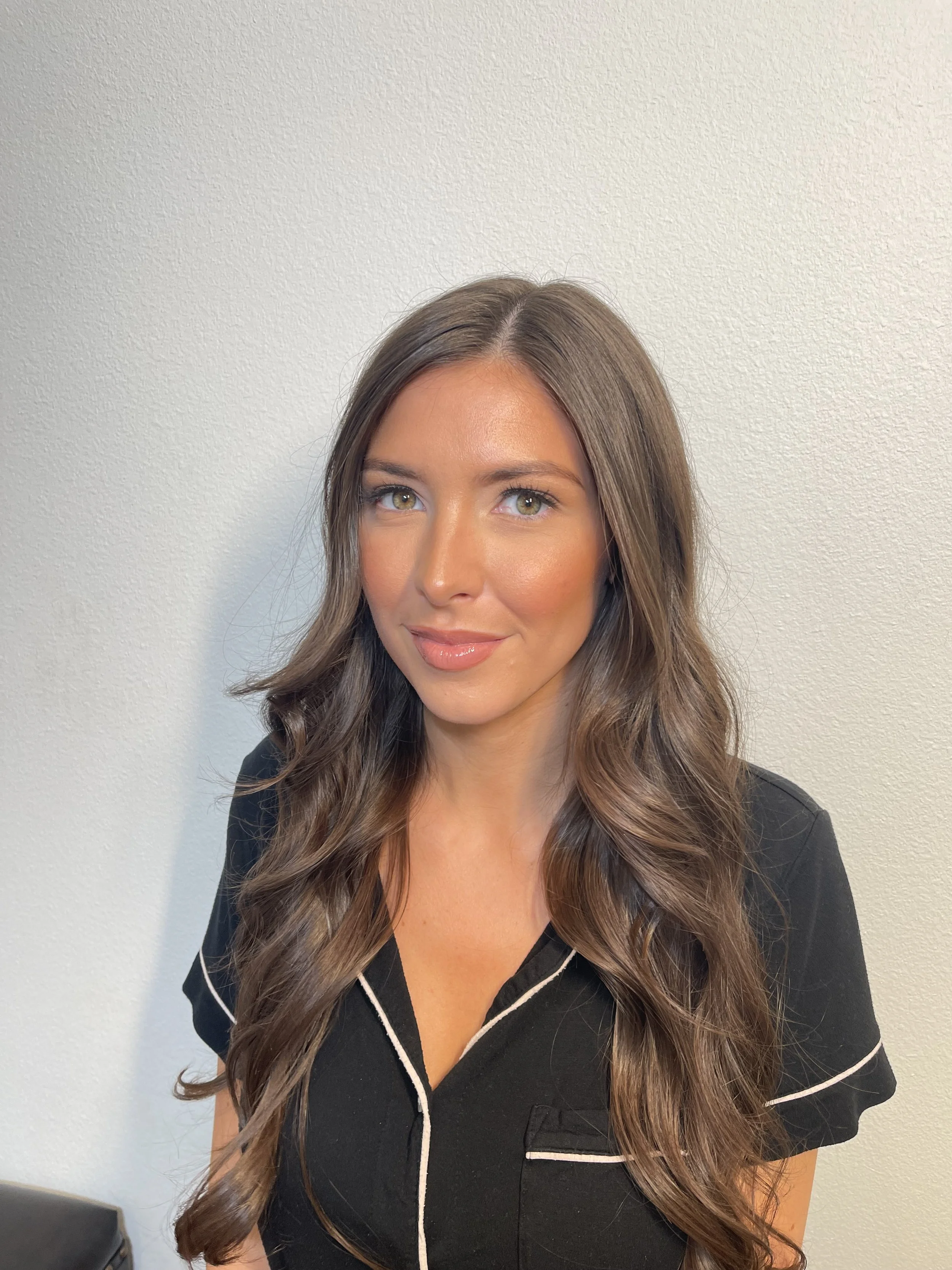 A woman with long wavy brown hair, tan skin, and green eyes is smiling softly and wearing a black top with white piping.