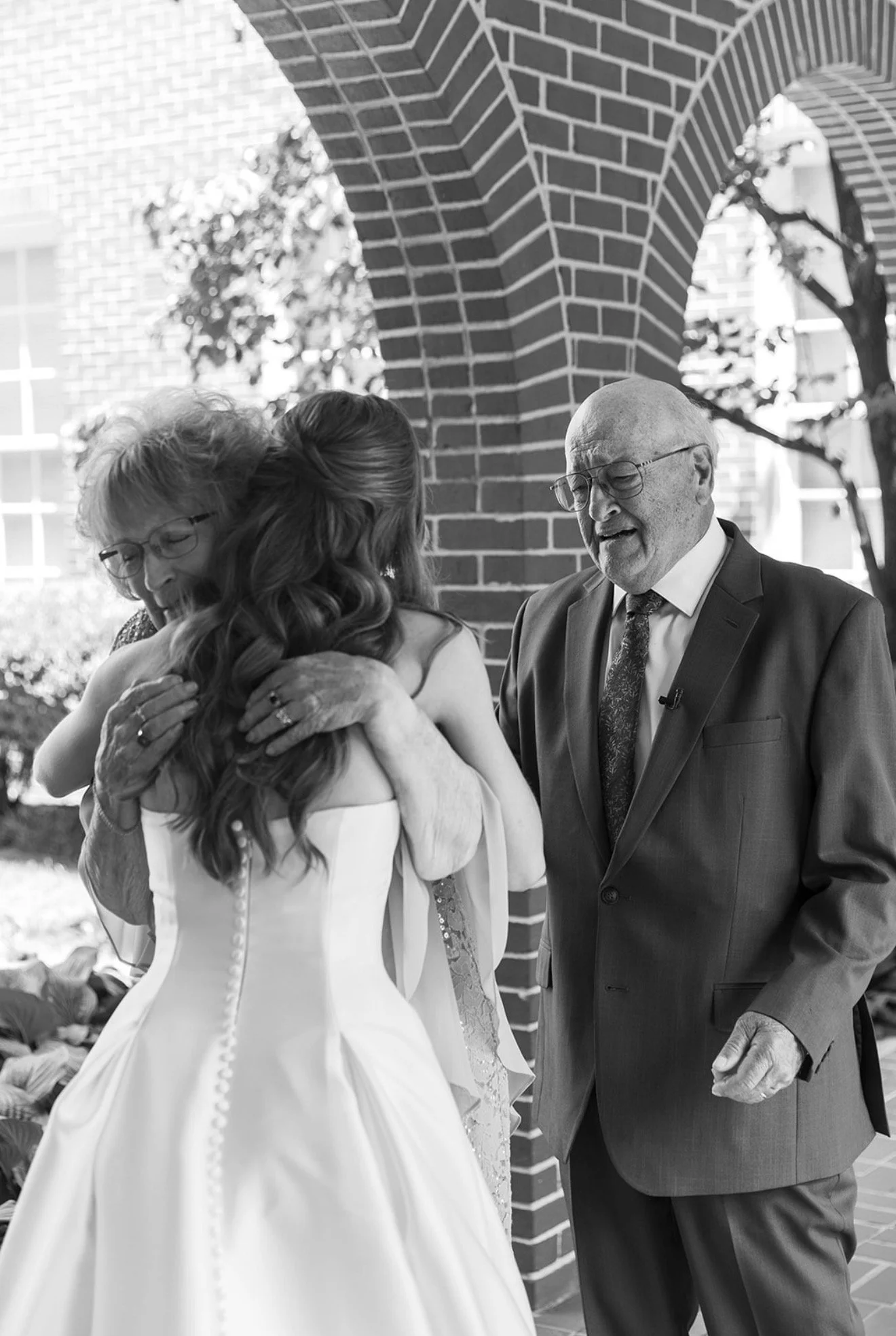 A bride hugging an elderly woman as another man looks on, outside on a sunny day near a brick building with arched openings.