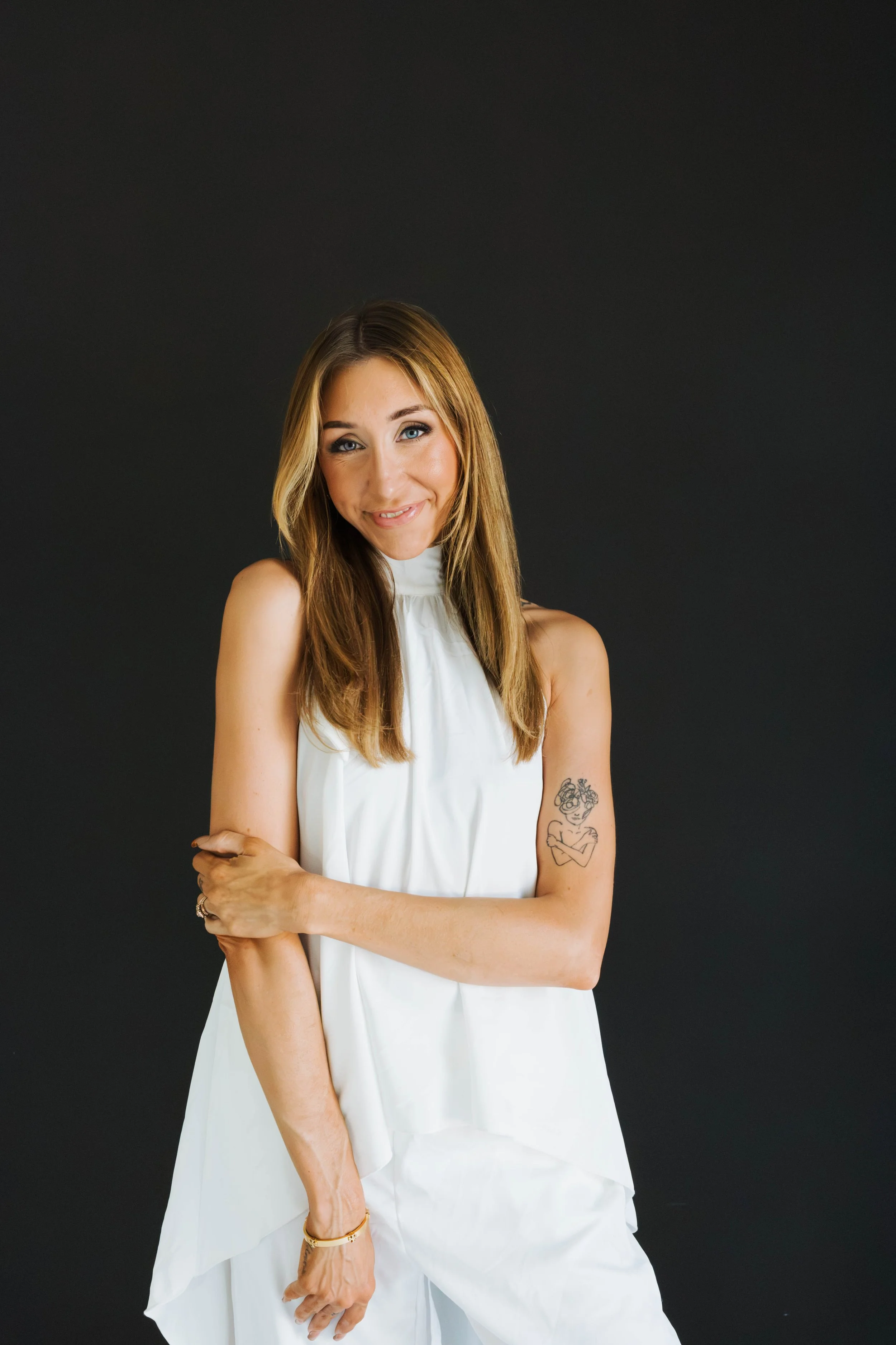 A young woman with long light brown hair, wearing a white sleeveless top and white pants, smiling and looking at the camera, standing against a plain dark background. She has tattoos on her left arm and wrist jewelry.