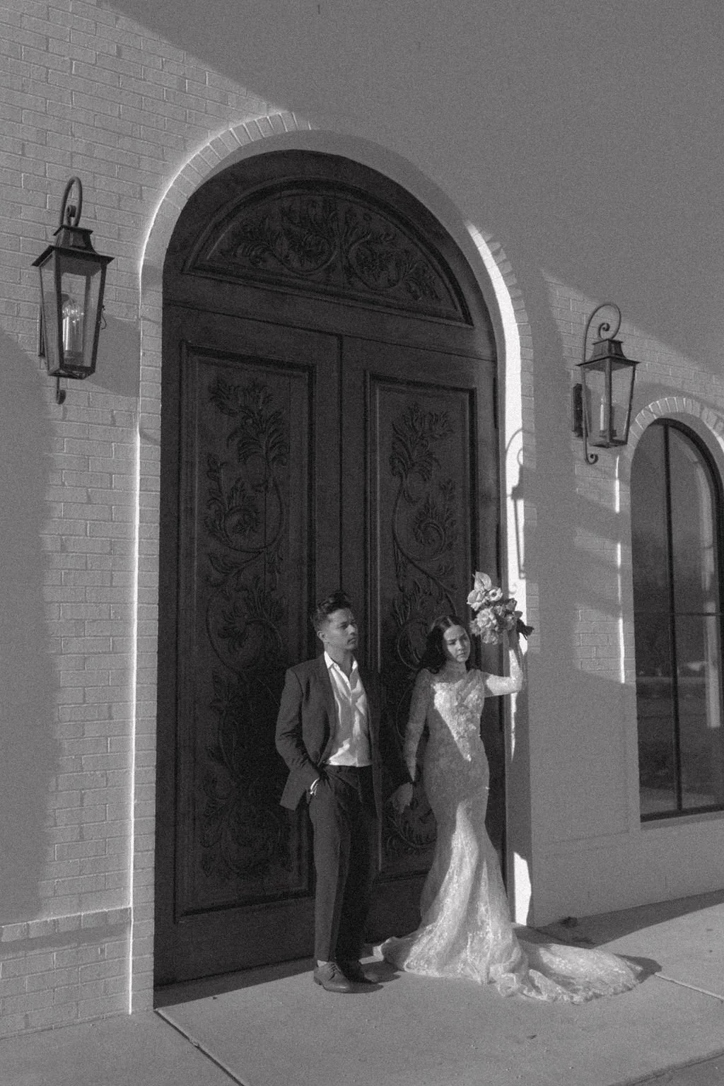 A black and white photo of a bride and groom standing outside a building with large wooden doors. The bride is holding a bouquet of flowers, wearing a lace wedding gown, and the groom is dressed in a suit.