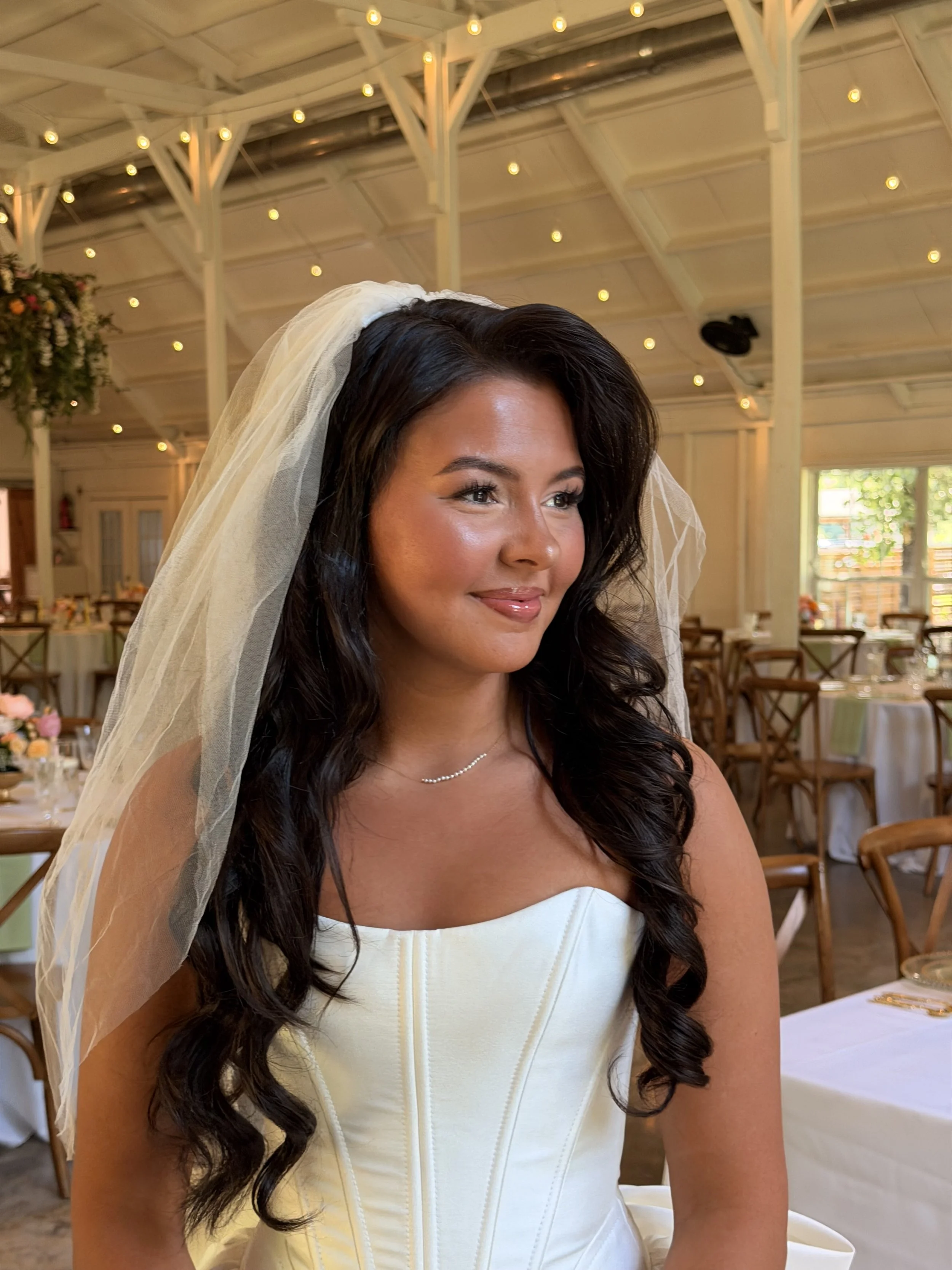 A smiling woman with long dark hair in a white strapless dress and a veil, sitting in a decorated reception hall with tables, chairs, and string lights.