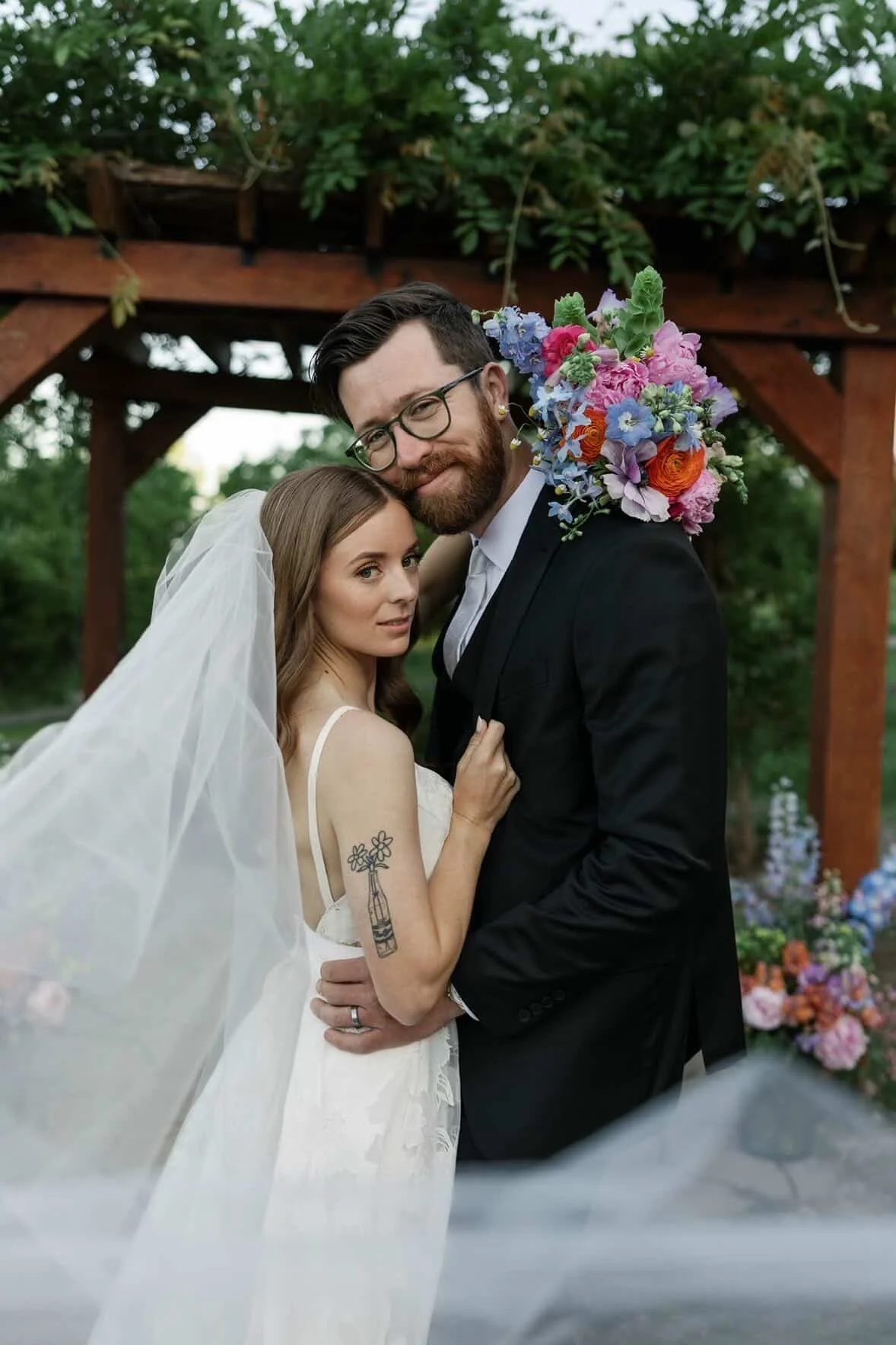 Bride and groom embracing outdoors beneath a wooden arch, with the bride wearing a wedding dress and veil, and the groom in a black suit, holding a colorful bouquet of flowers on his shoulder.