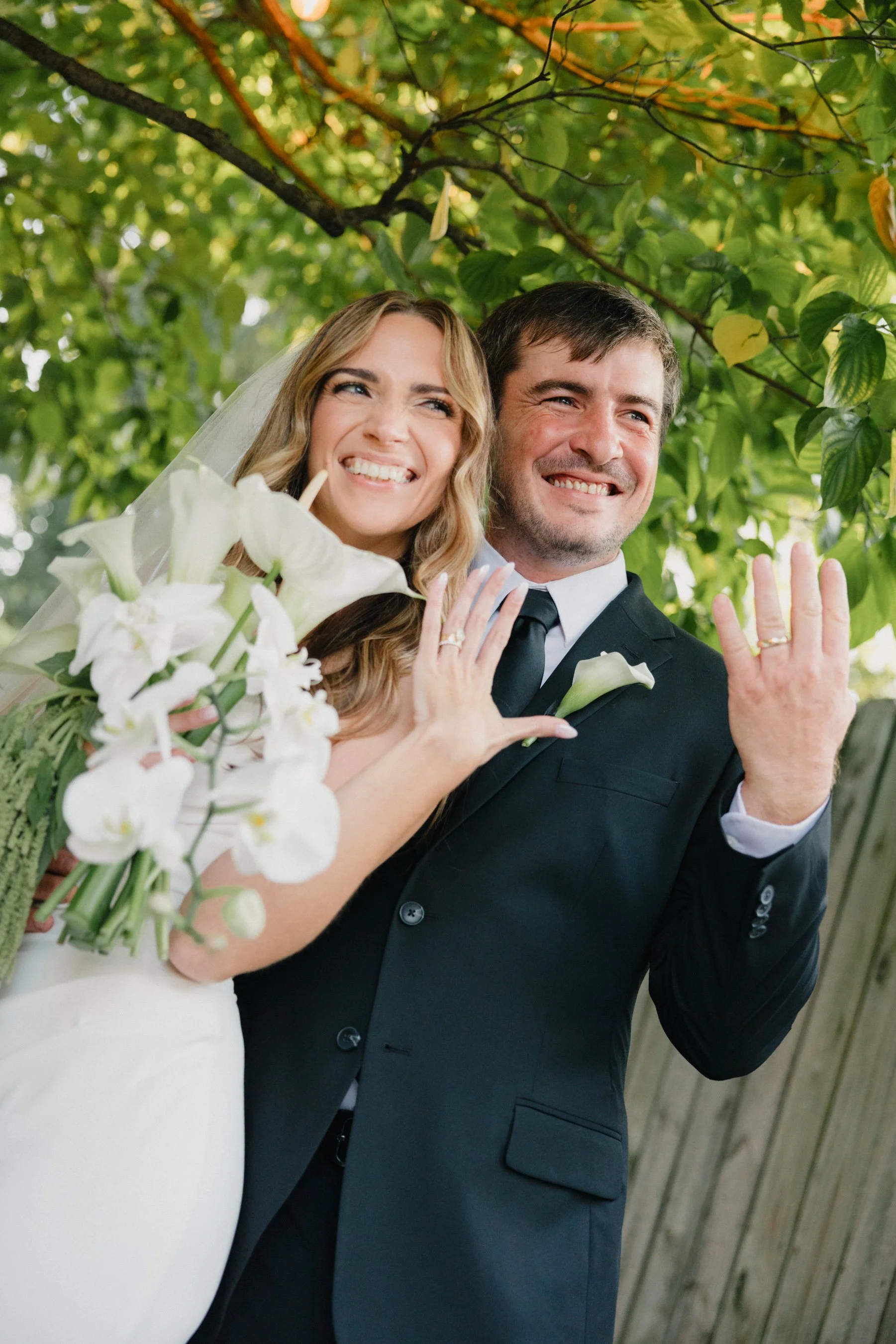 A newlywed couple smiling and showing off their wedding rings outdoors under a tree, with green leaves and sunlight in the background. The bride holds a bouquet of white flowers.