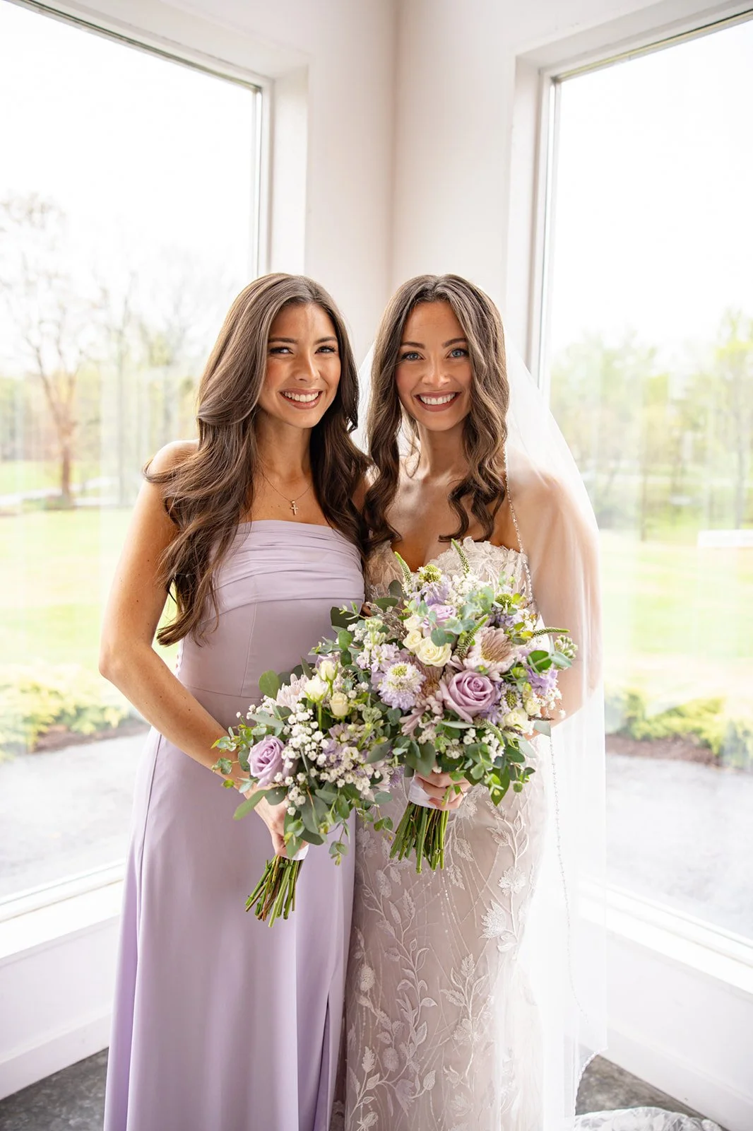 Two women, one in a lavender dress and the other in a wedding gown, smiling and holding bouquets, standing inside by large windows with a view of a garden outdoor.