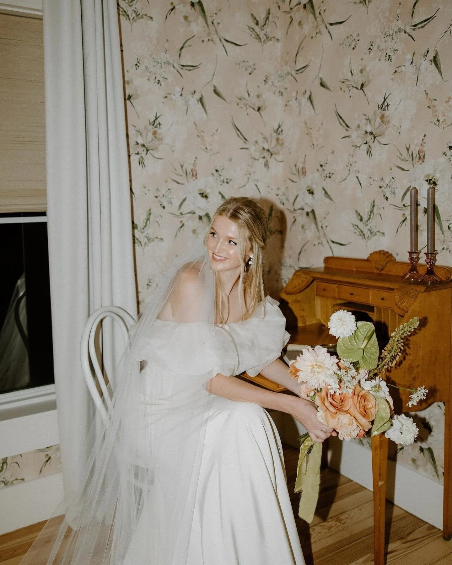 A woman with long blonde hair styled in loose waves sits on a brown leather chair in a room with white walls and wooden furniture, looking to her left with a slight smile.