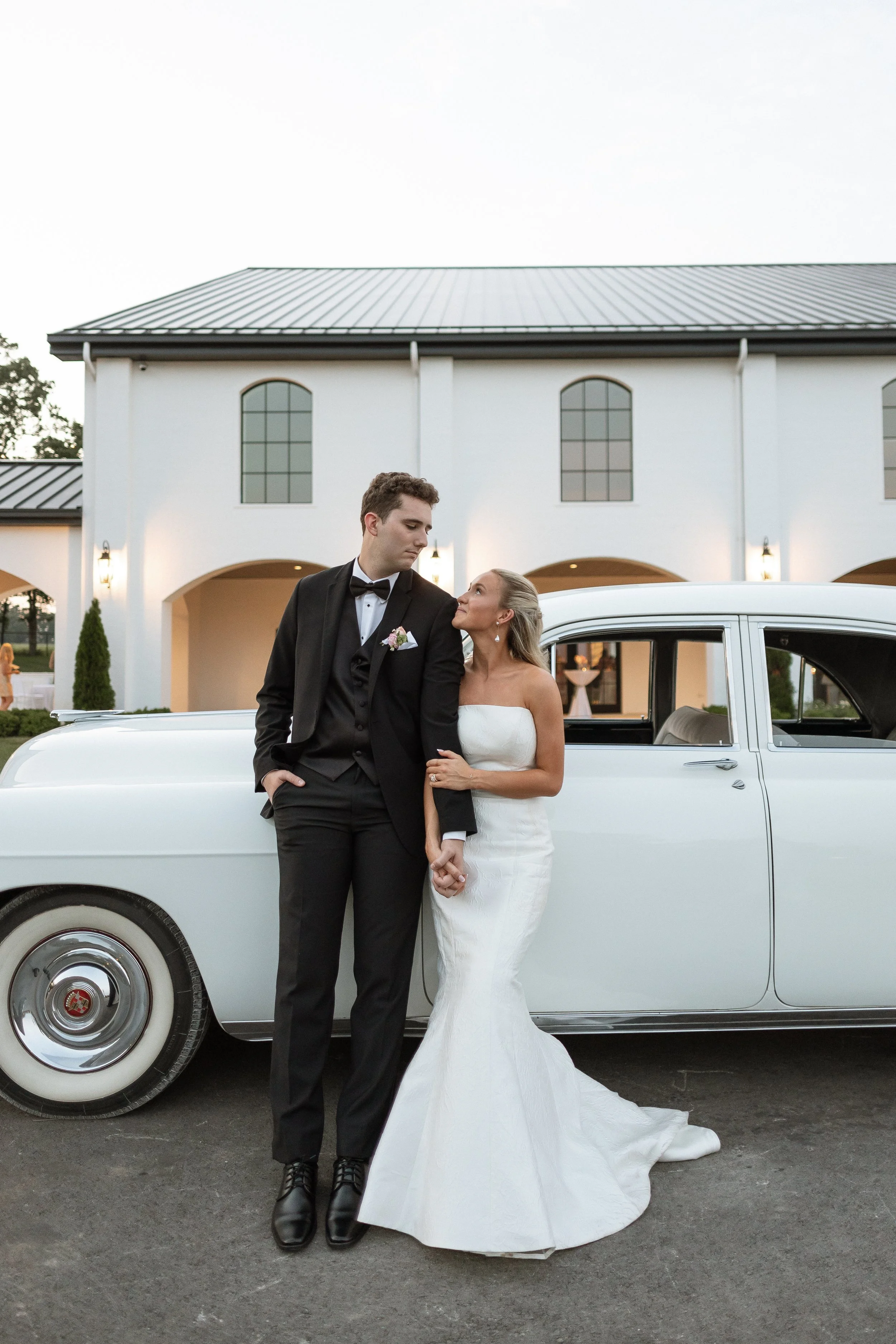 A bride and groom stand in front of a vintage white car outside a white building during their wedding. The bride is wearing a strapless white wedding dress, and the groom is dressed in a black tuxedo with a bow tie.