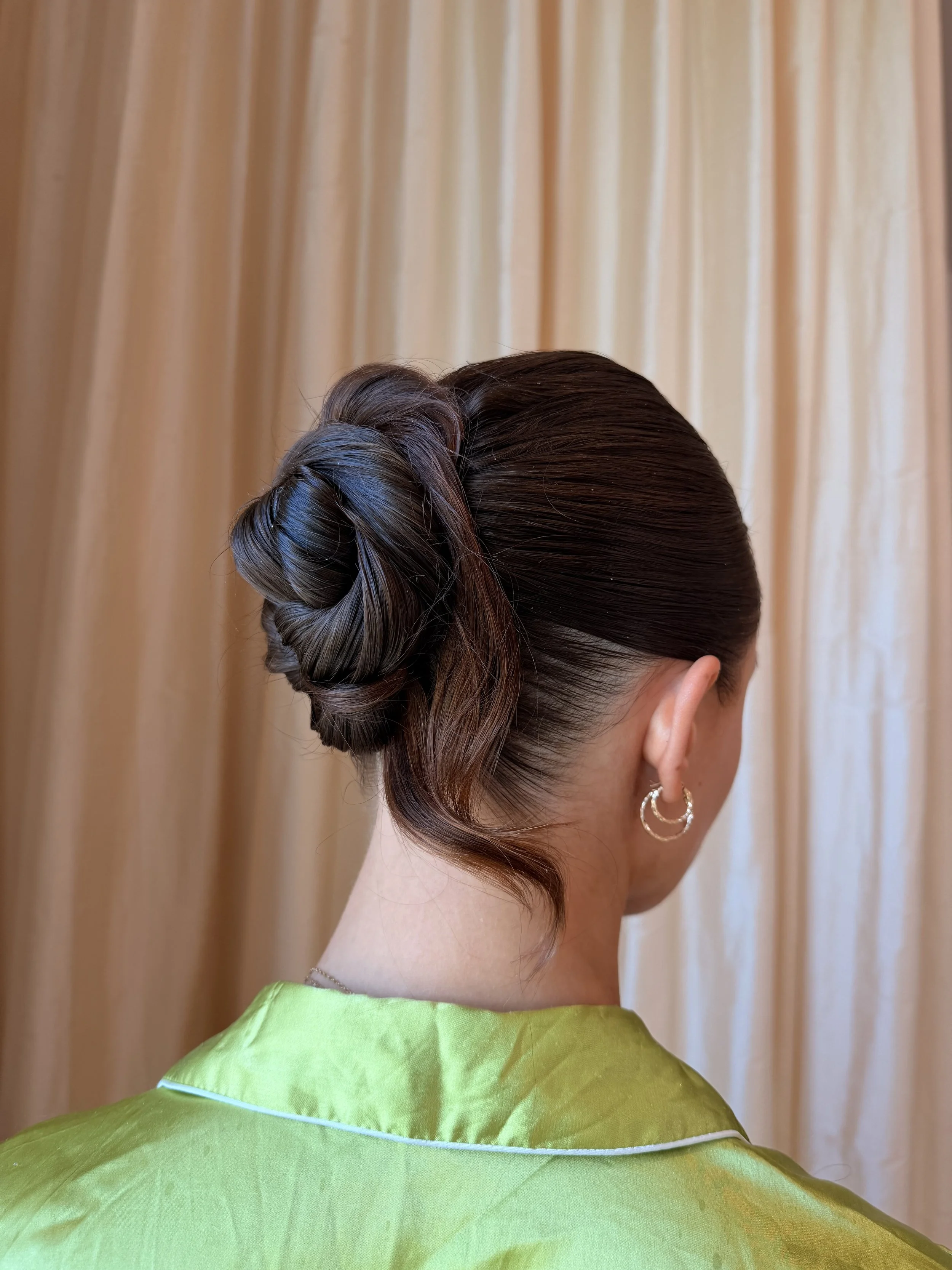 Rear view of a woman with dark brown hair styled in an elegant updo, wearing hoop earrings, a green traditional dress, and standing in front of a beige curtain.