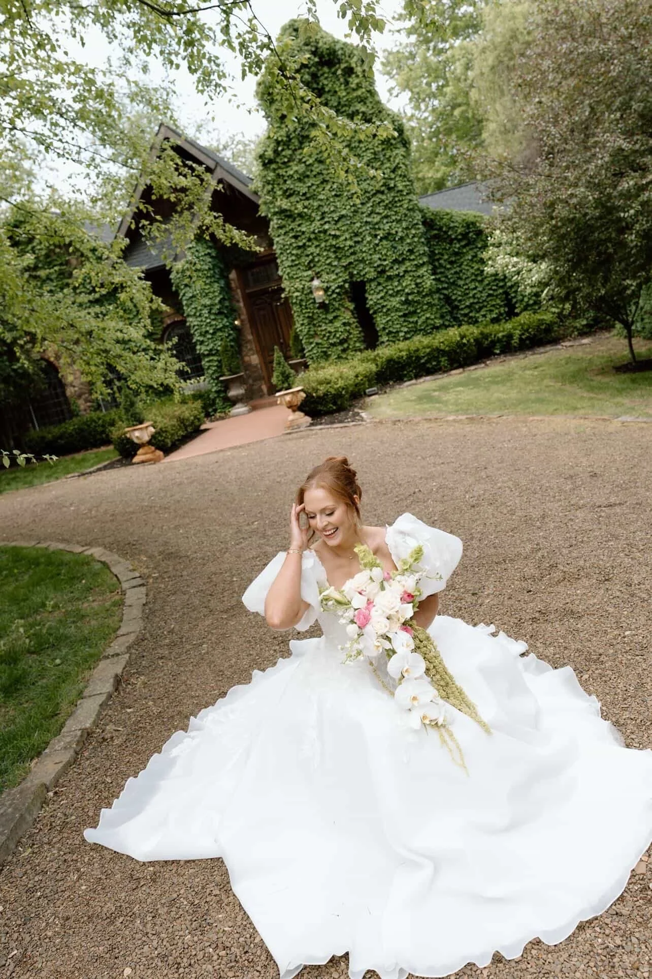A woman dressed in a white wedding gown is sitting outdoors on a gravel path, holding a bouquet of flowers, and smiling while touching her hair, in front of a large house covered with green ivy and surrounded by trees.