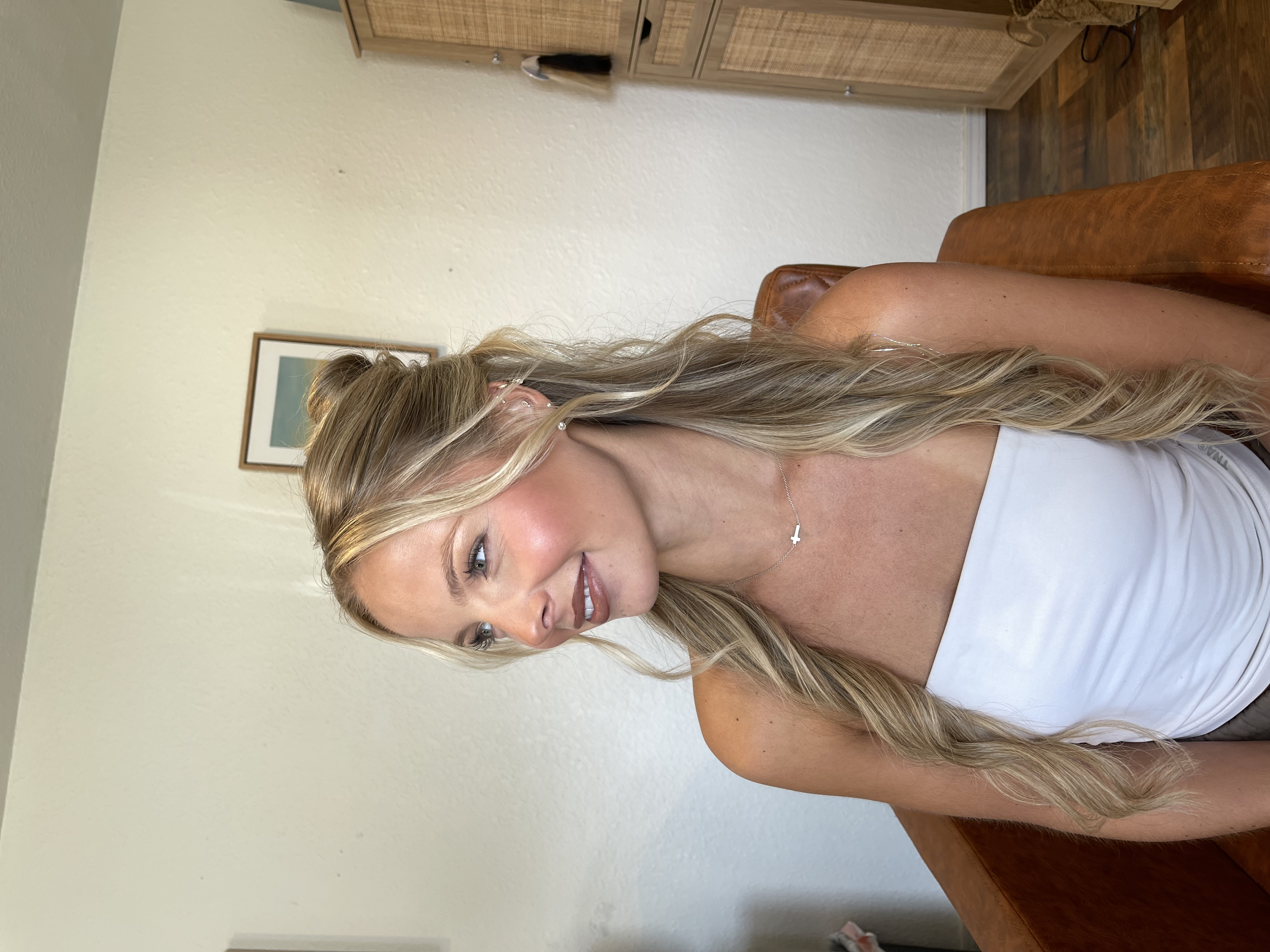 A woman with long, wavy blonde hair sitting on a brown leather chair in a room with beige walls, a wooden dresser, and a framed picture on the wall.