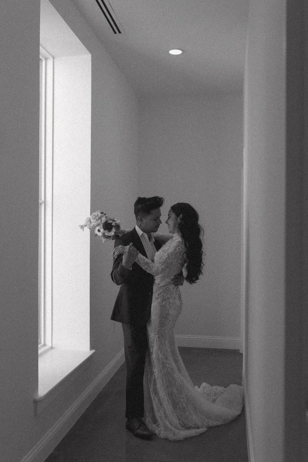 A black and white photo of a couple dancing, the woman in a lace wedding dress and the man in a suit, in a simple room with a window and a small bouquet of flowers.