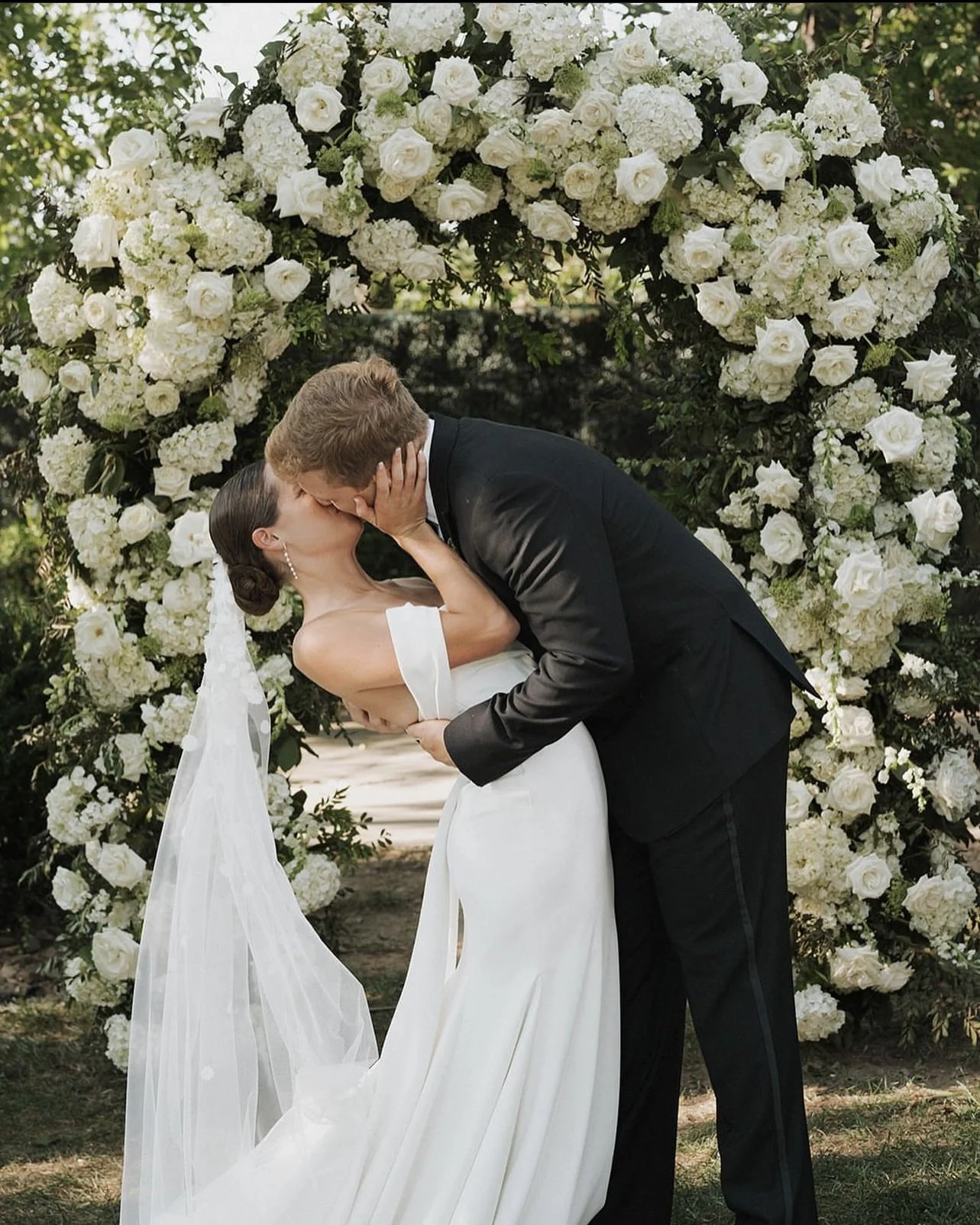 A bride and groom kissing under an arch of white flowers during their wedding ceremony.