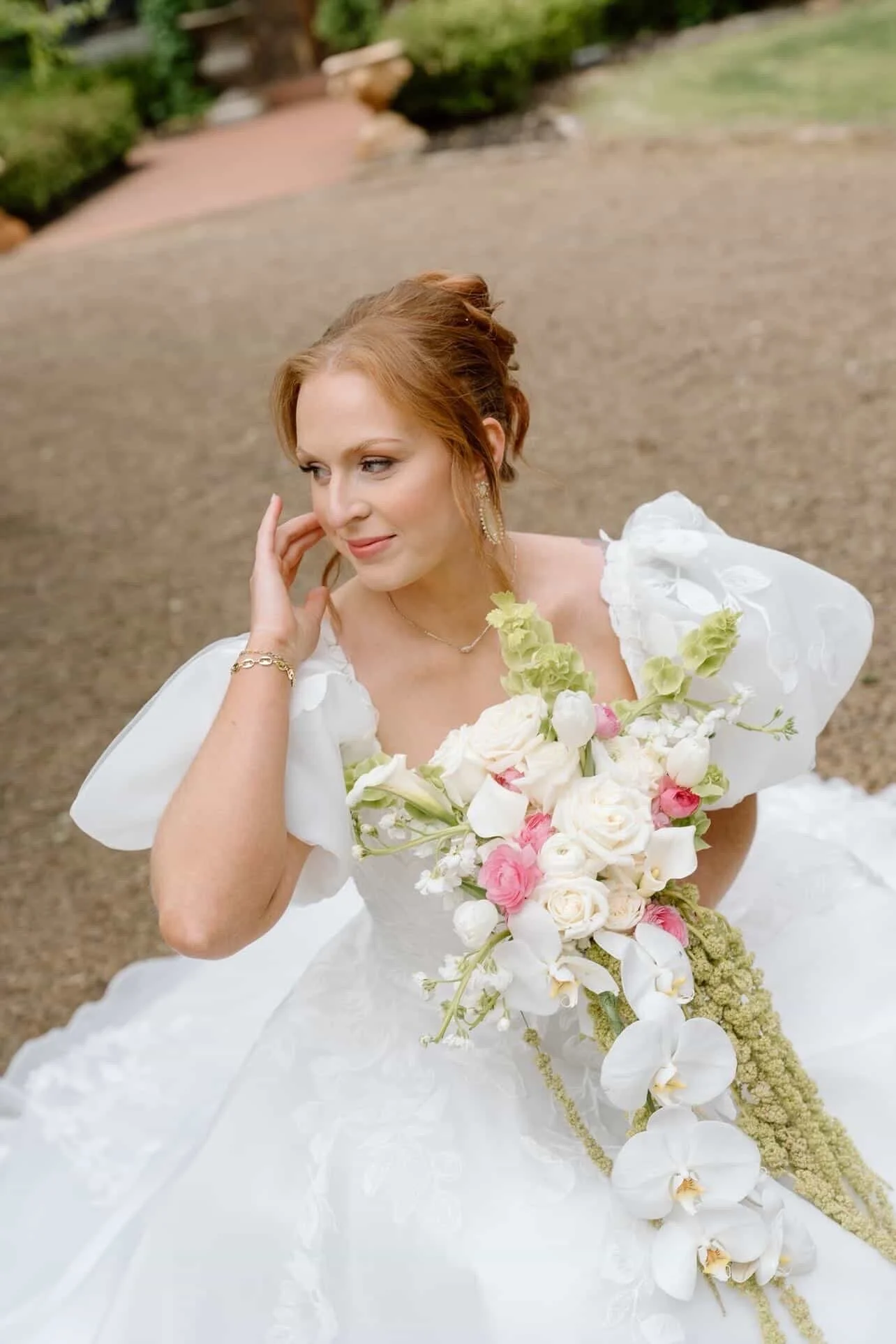 A bride with red hair styled in an elegant updo, wearing a white wedding dress with puffed sleeves and lace details, holding a cascading bouquet of white, pink, and cream flowers, with greenery and orchids, standing outdoors in a garden setting.